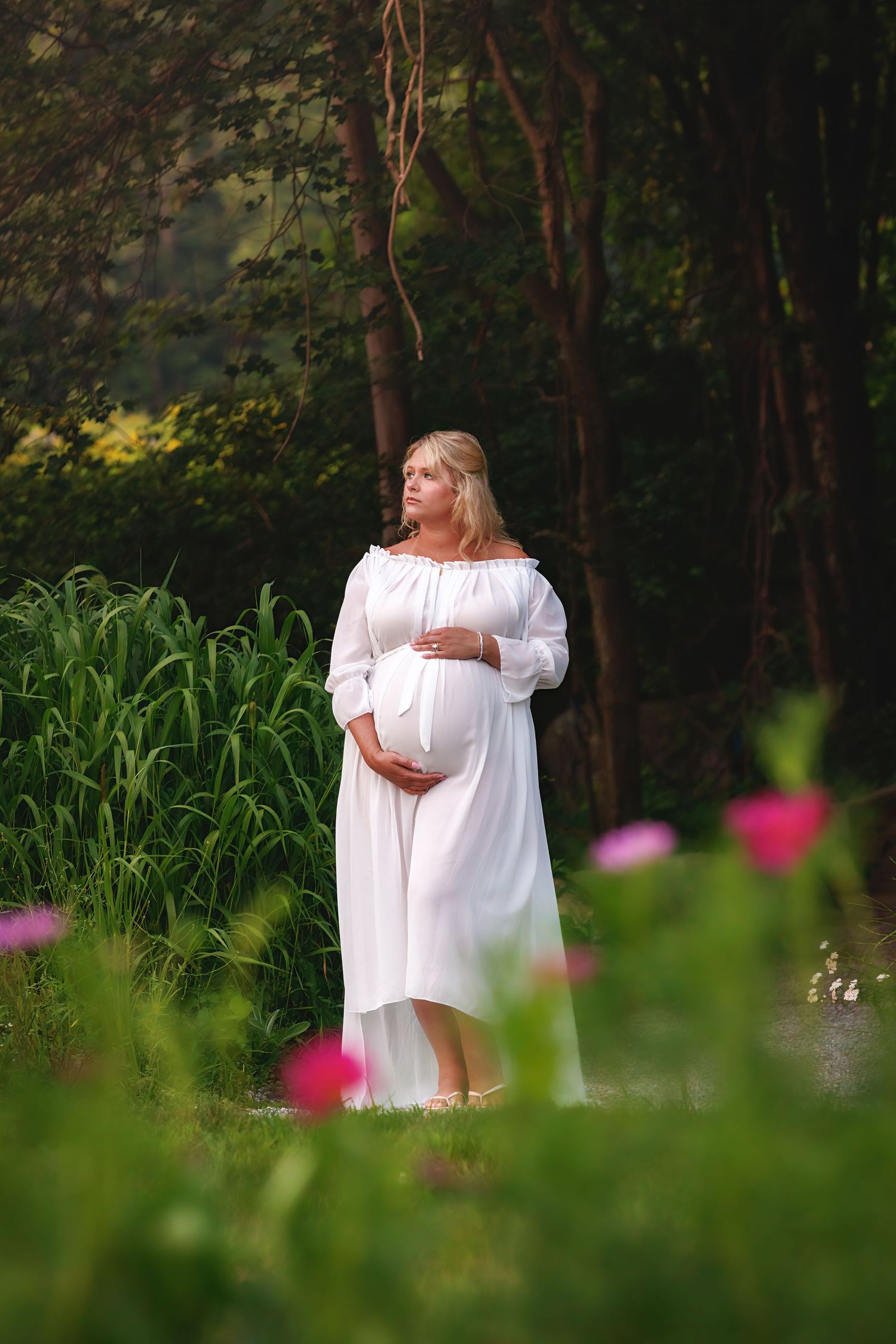 A figure in a flowing white dress stands in a serene woodland setting with green foliage and pink flowers in the foreground.