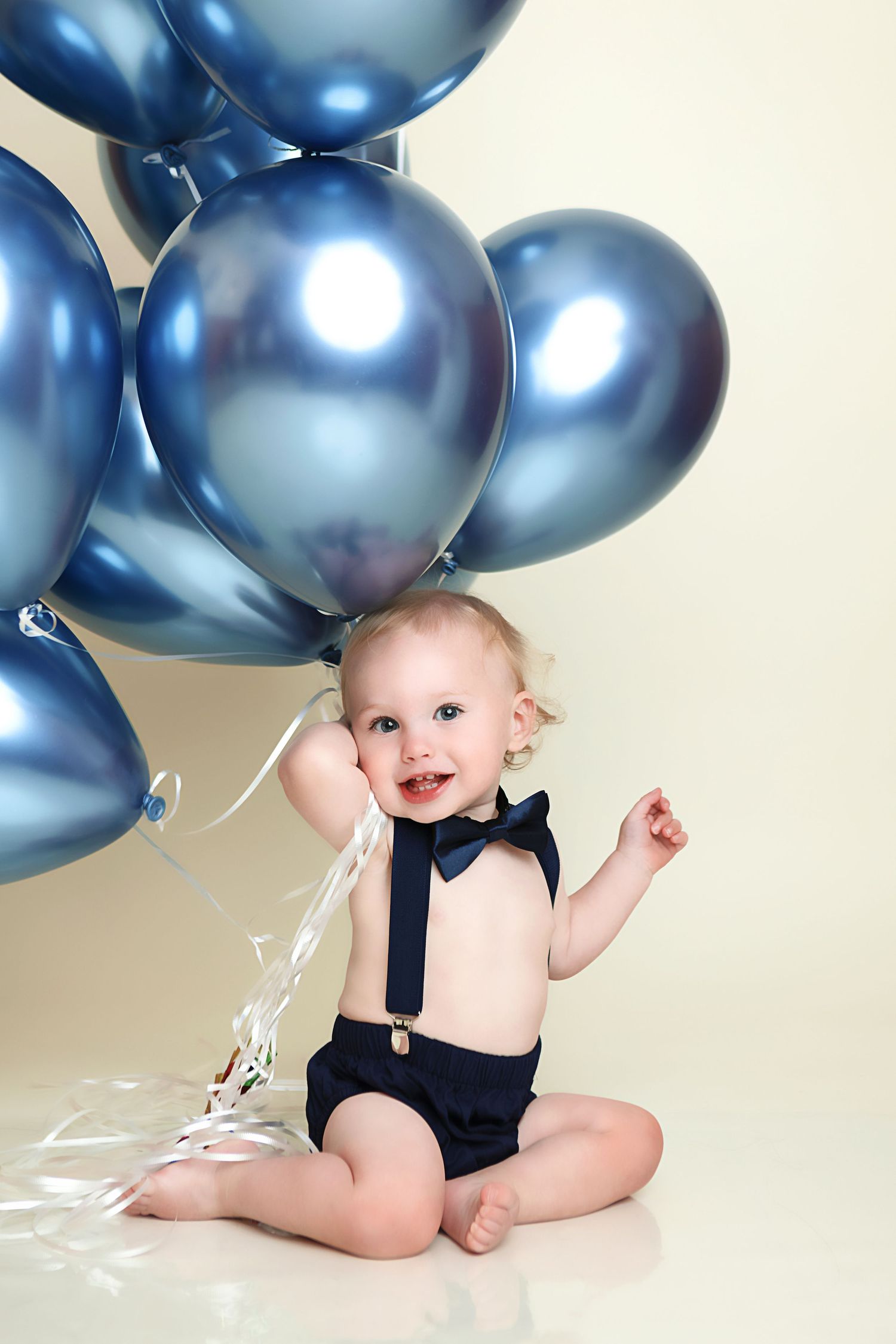 Baby in black bow tie and suspenders poses with metallic blue balloons during first birthday photo session.