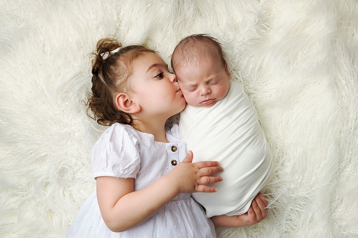A toddler in white clothing gives a gentle kiss to a sleeping baby on white bedding.