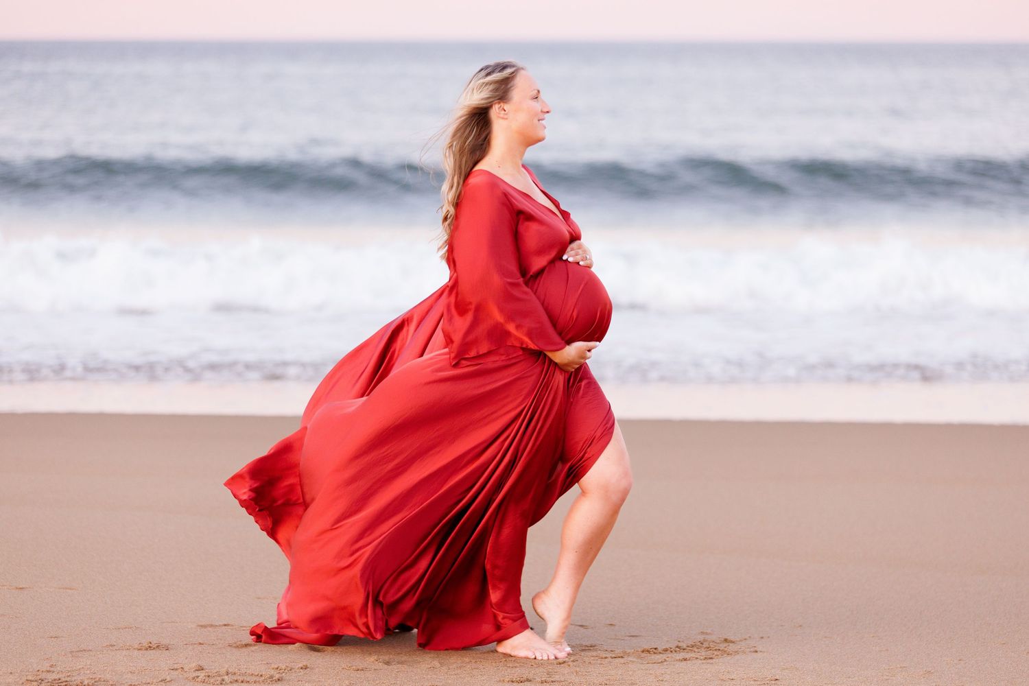Pregnant woman in flowing red dress holding baby bump on sandy beach with ocean waves backdrop. Luxury maternity photography near Nashua NH