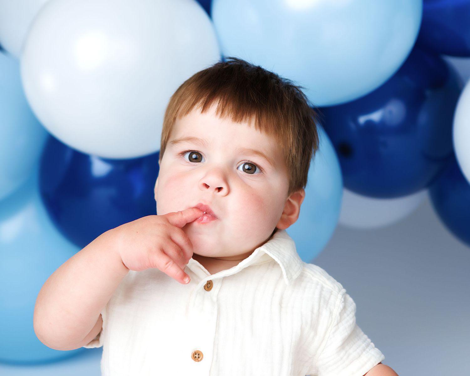 Toddler boy in white shirt with finger in mouth at first birthday party with blue and white balloons.