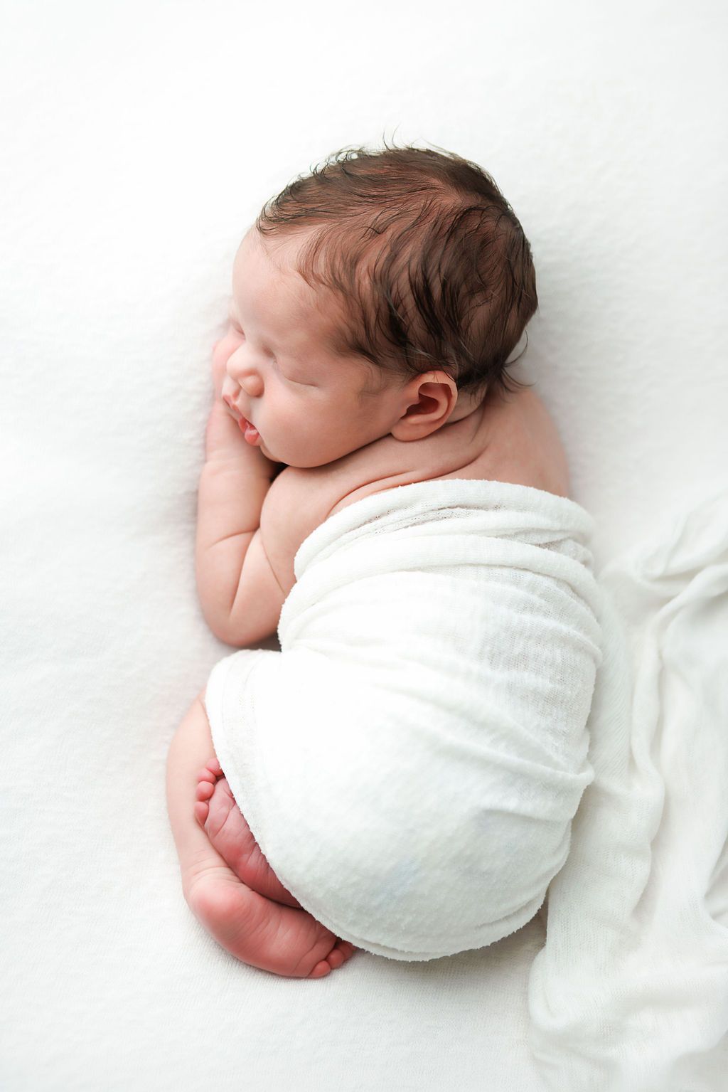 Peaceful newborn baby sleeping curled up, wrapped in a soft white swaddle blanket on a white surface.