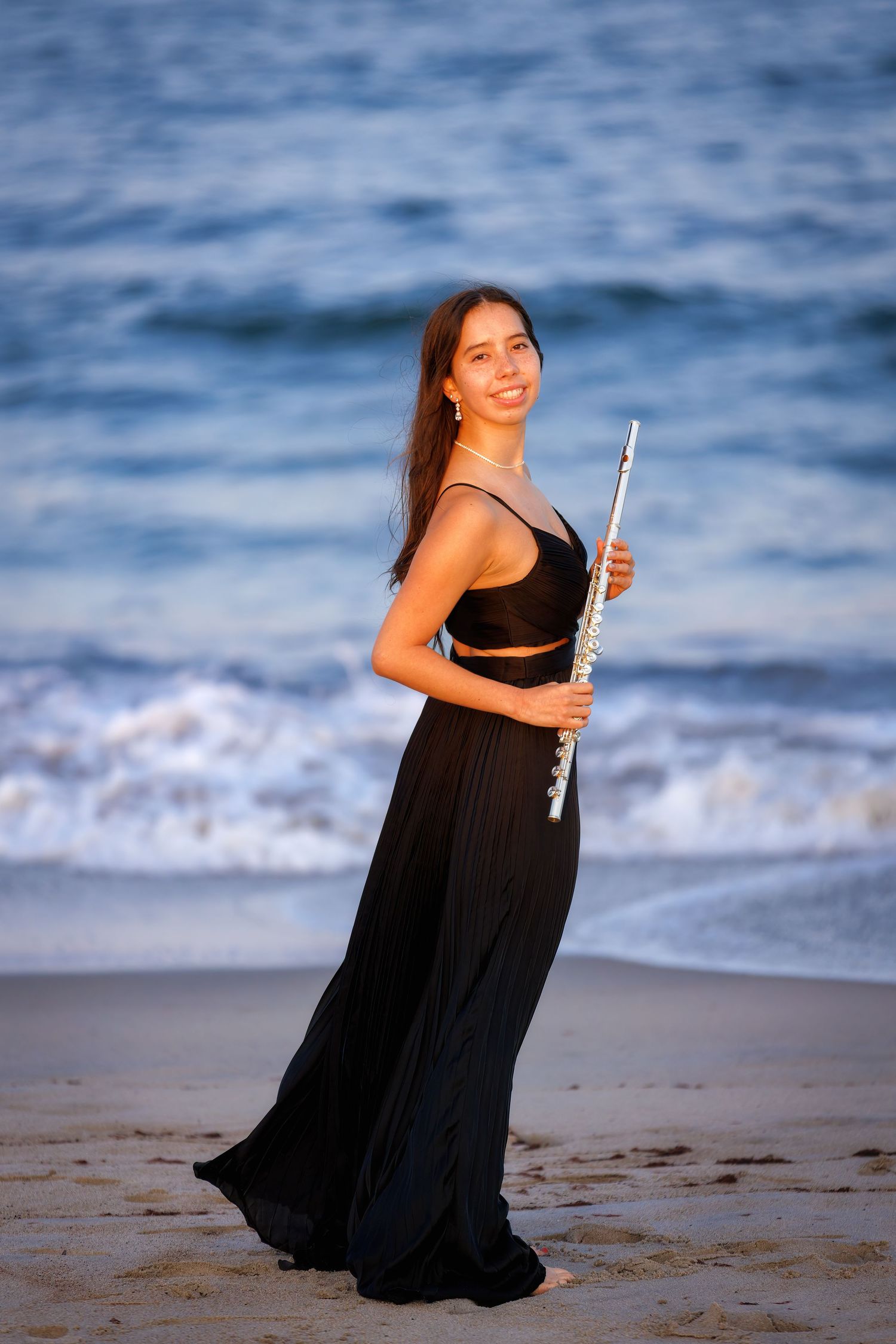 Beach Senior Portrait. A musician in a flowing black dress poses with a flute on the beach at sunset.