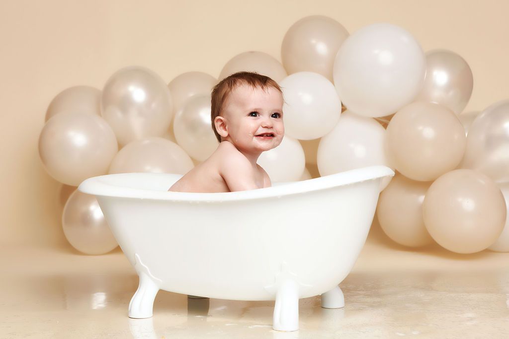 Smiling baby sitting in a white mini bathtub surrounded by cream and white balloons on beige background.