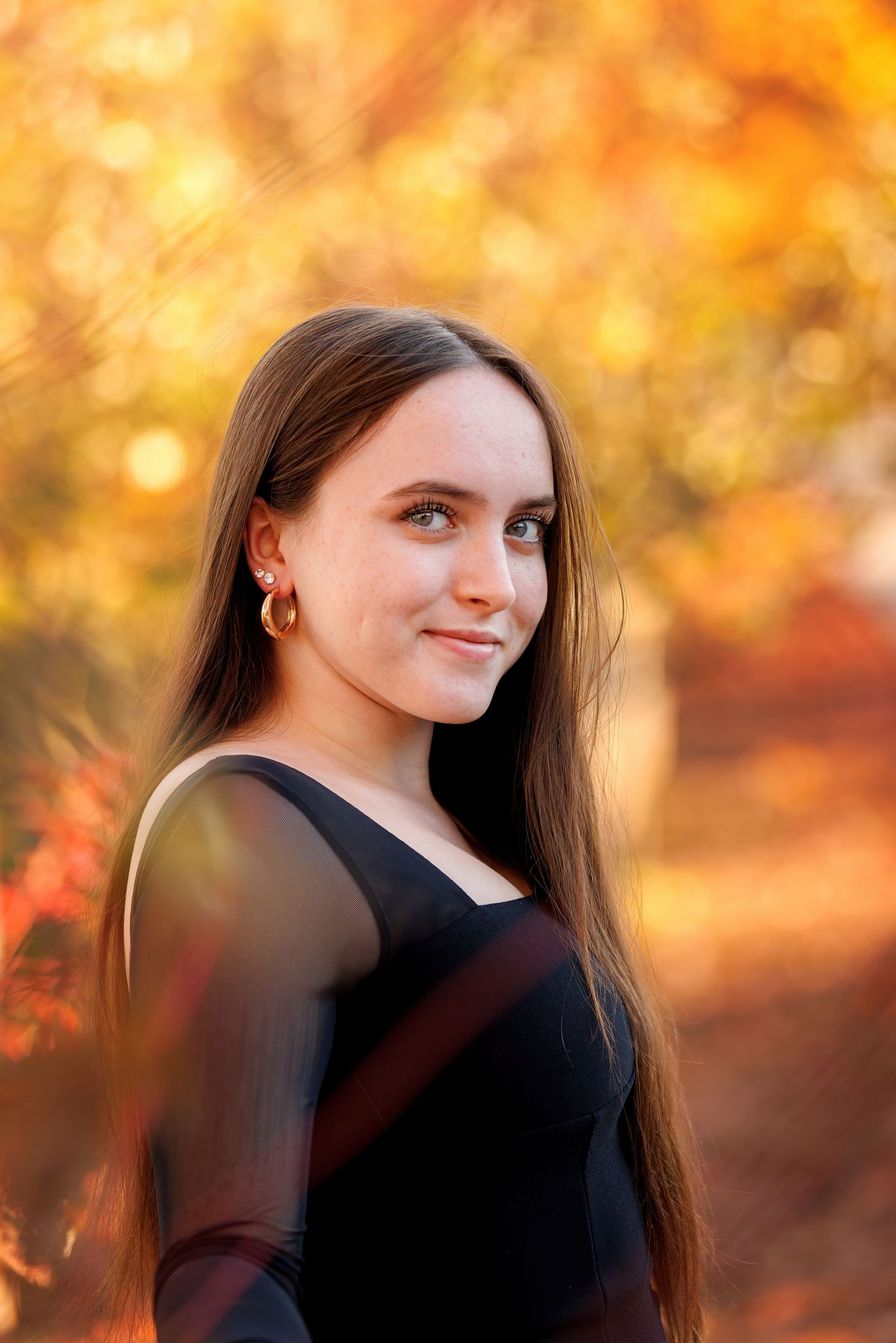 Young woman with long brown hair wearing a black dress poses outdoors among vibrant autumn foliage.