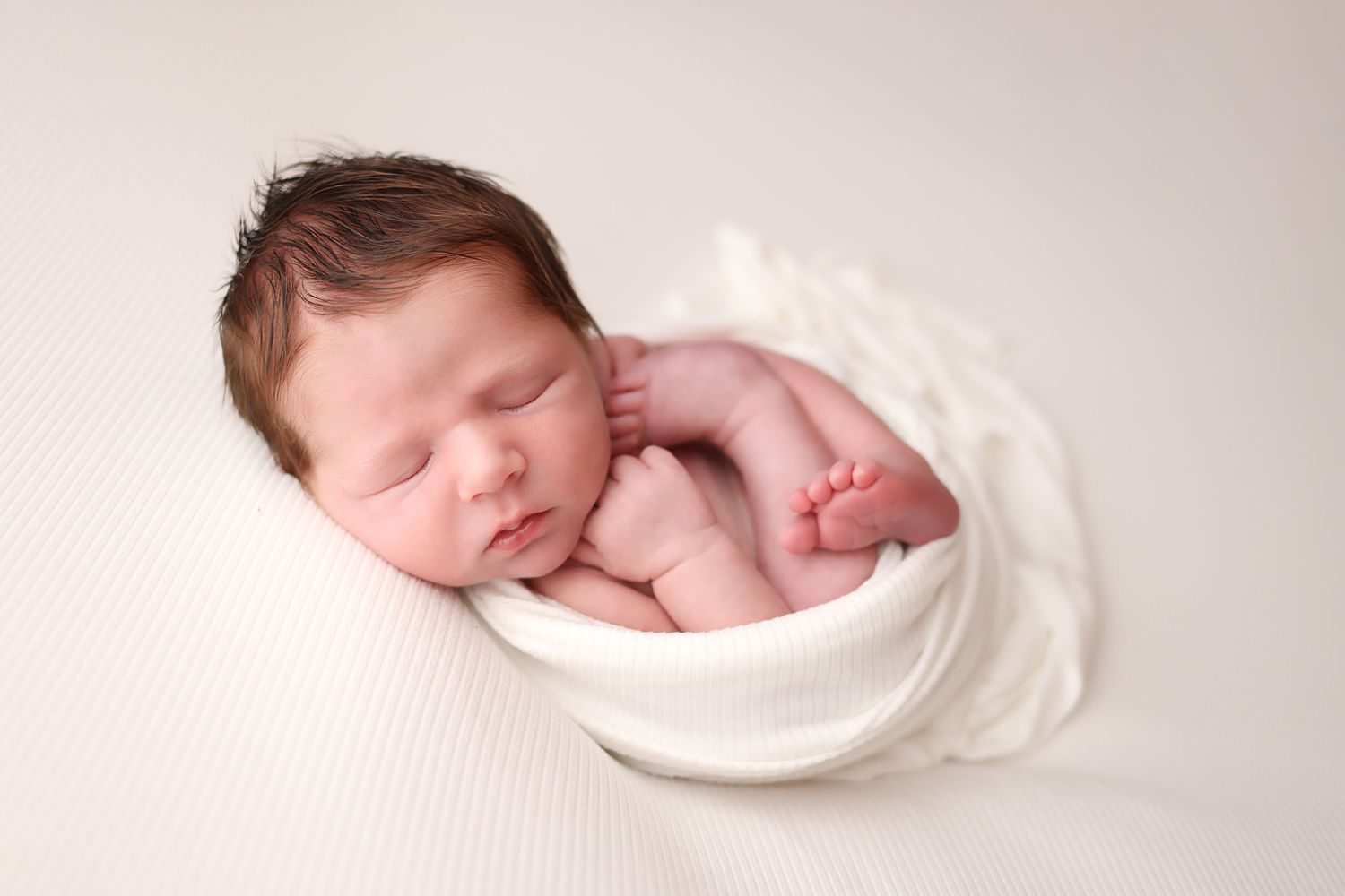 A sleeping newborn baby wrapped in soft white fabric lies peacefully against a light backdrop.