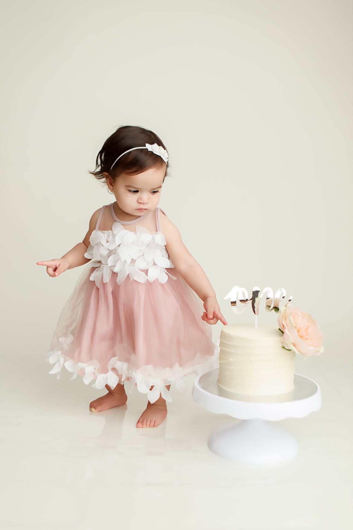 Baby in pink tulle dress with white flowers reaching for elegant white birthday cake on light background.