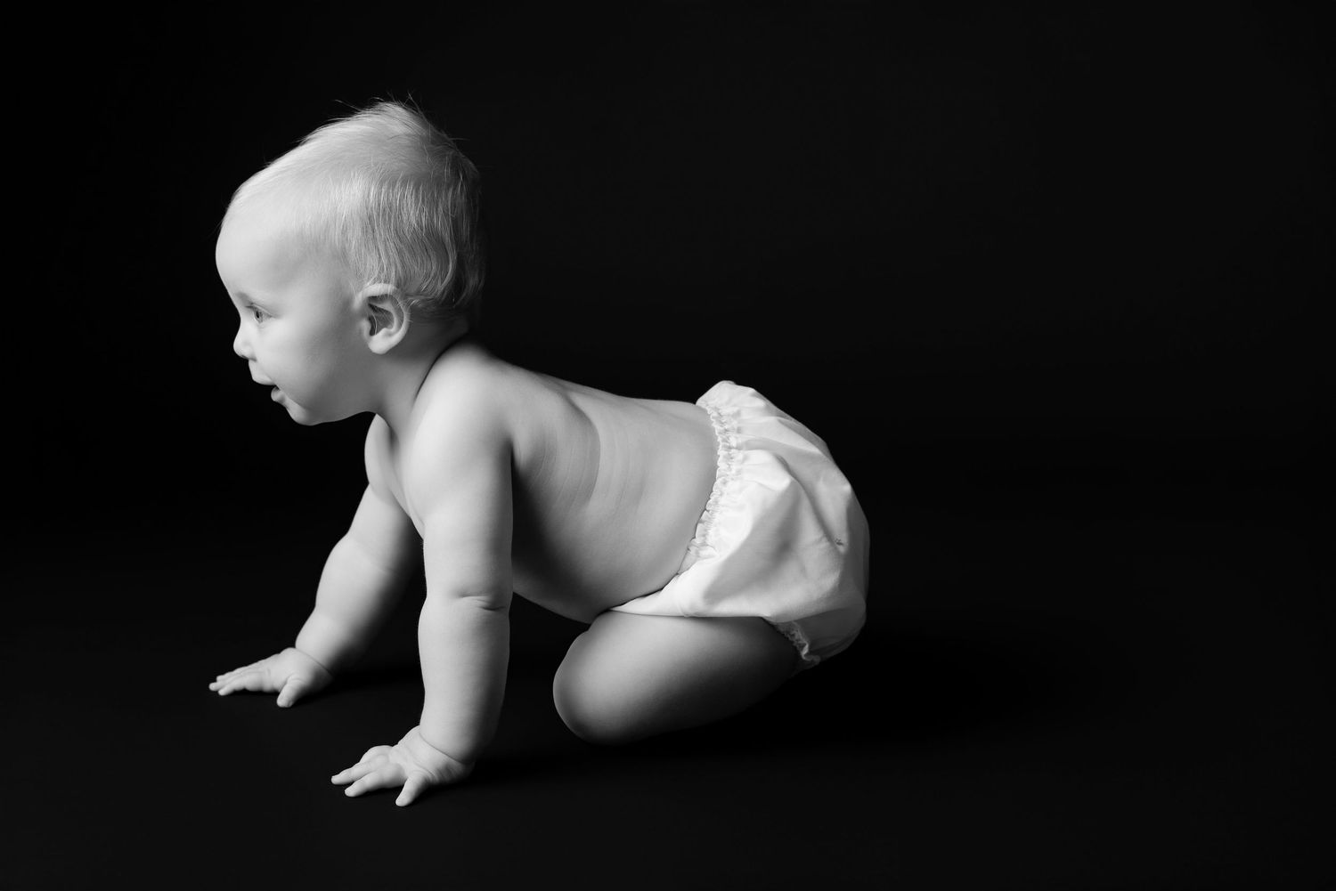 Black and white photo of a baby crawling in a diaper against a dark background.