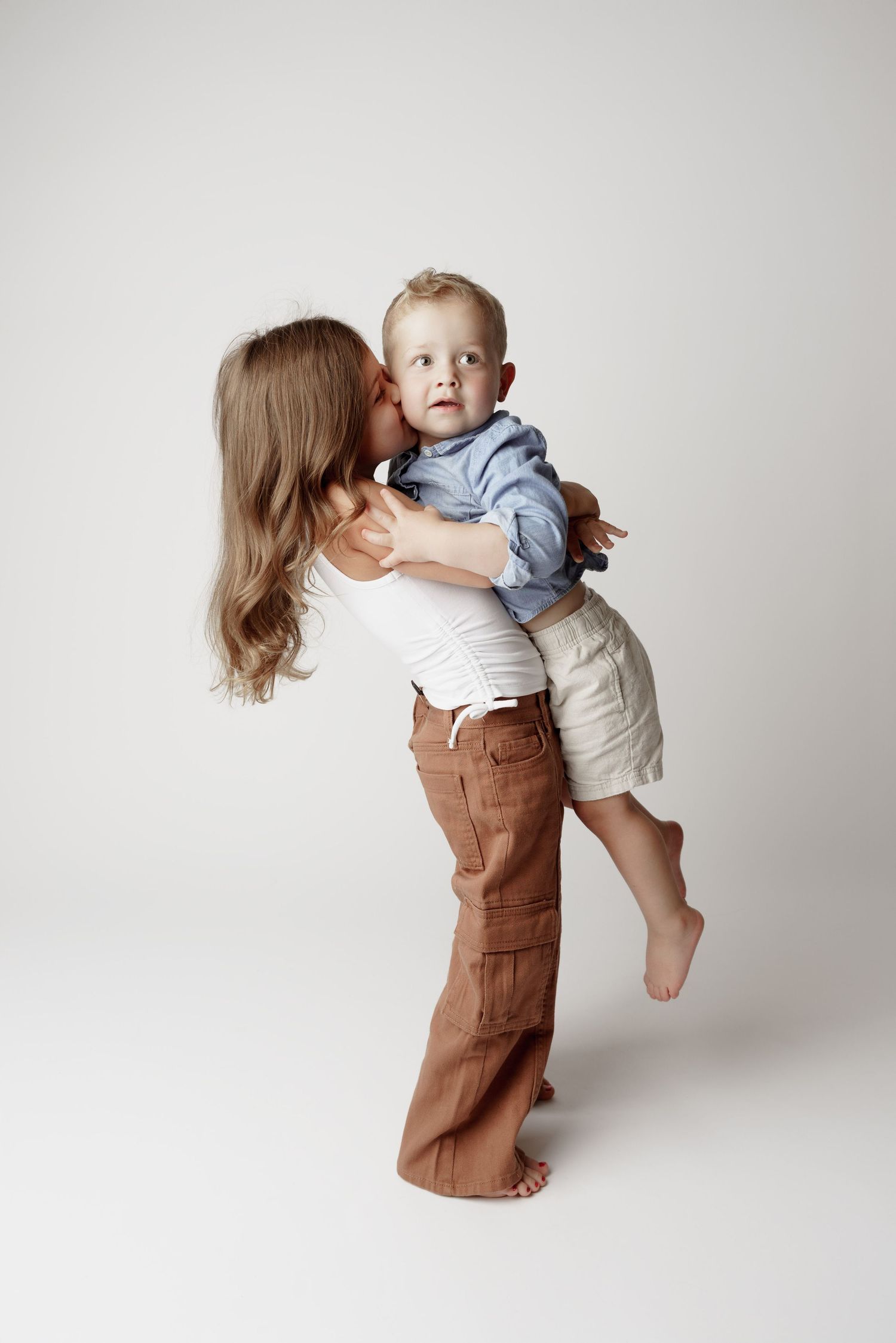 Young girl in white top and brown pants hugging and kissing a toddler boy in a blue shirt.