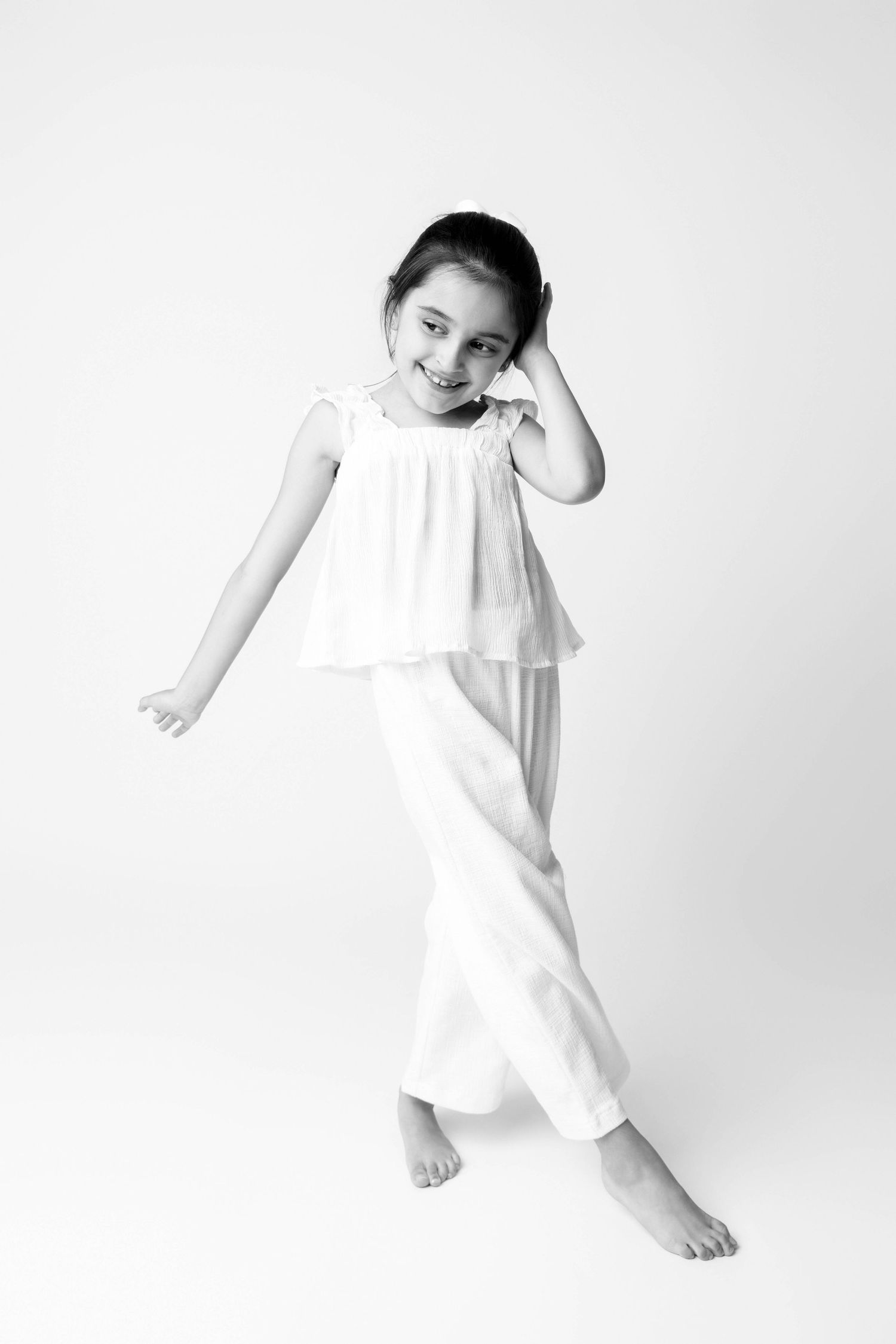 A cheerful young girl in white outfit poses barefoot, smiling in a black and white studio portrait.