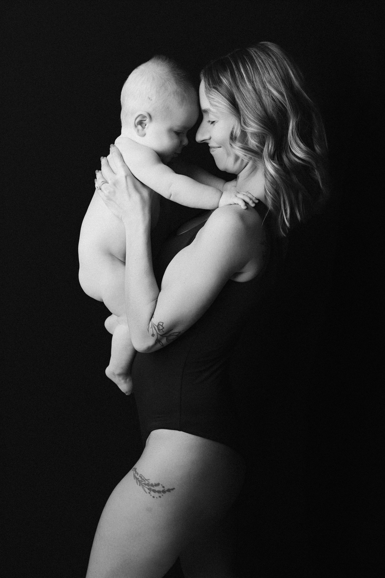 A tender black and white portrait of a parent and baby sharing an intimate moment against a dark background.