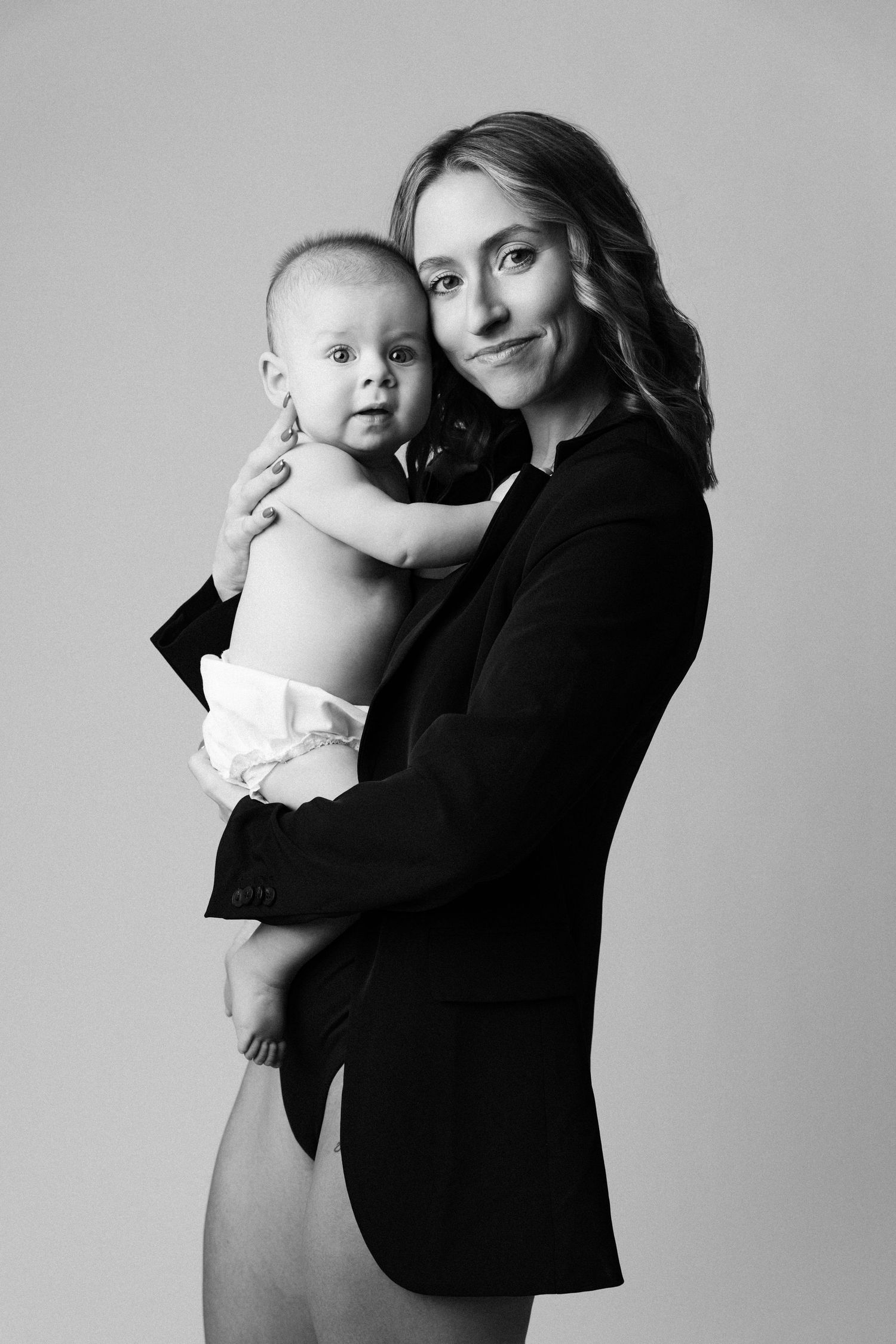 A black and white portrait of a parent in a black blazer holding a baby in diapers against a light background.