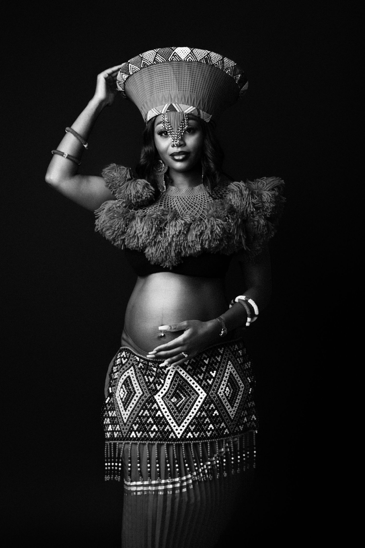 Black and white photo of a pregnant African woman in traditional beaded attire, holding a woven basket on her head.