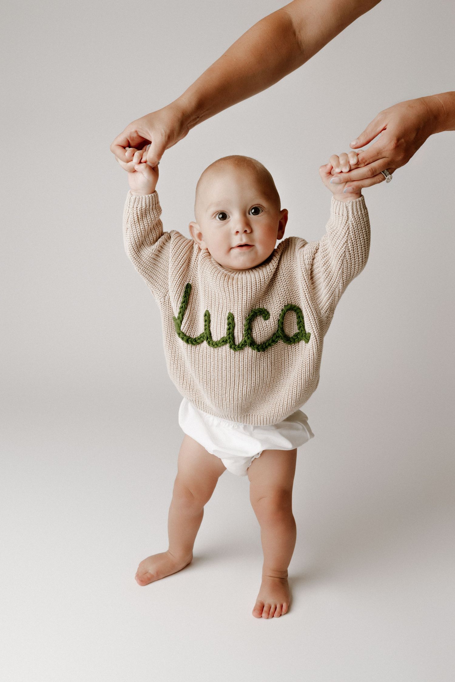 Baby wearing a beige knit sweater with green 'luca' lettering, held upright by adult hands on white background.