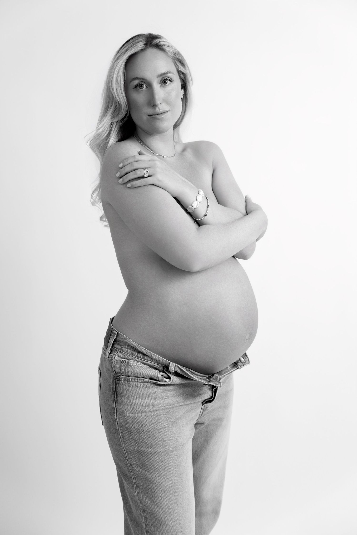 Black and white artistic maternity portraits showing a woman in jeans embracing her pregnant belly against white background.