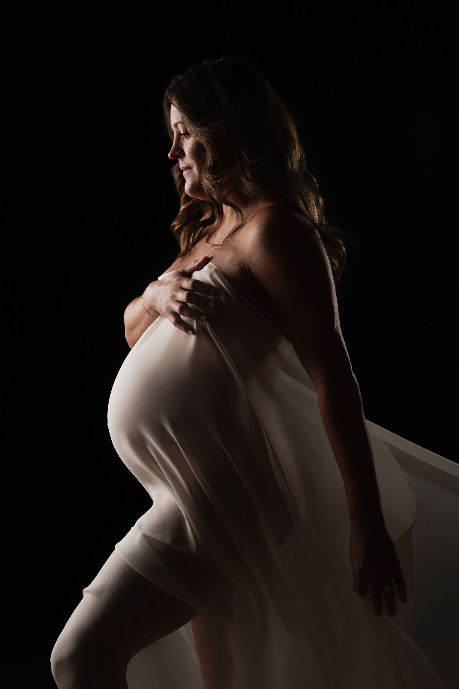Pregnant woman draped in white fabric poses gracefully against a black background in a studio maternity portrait.