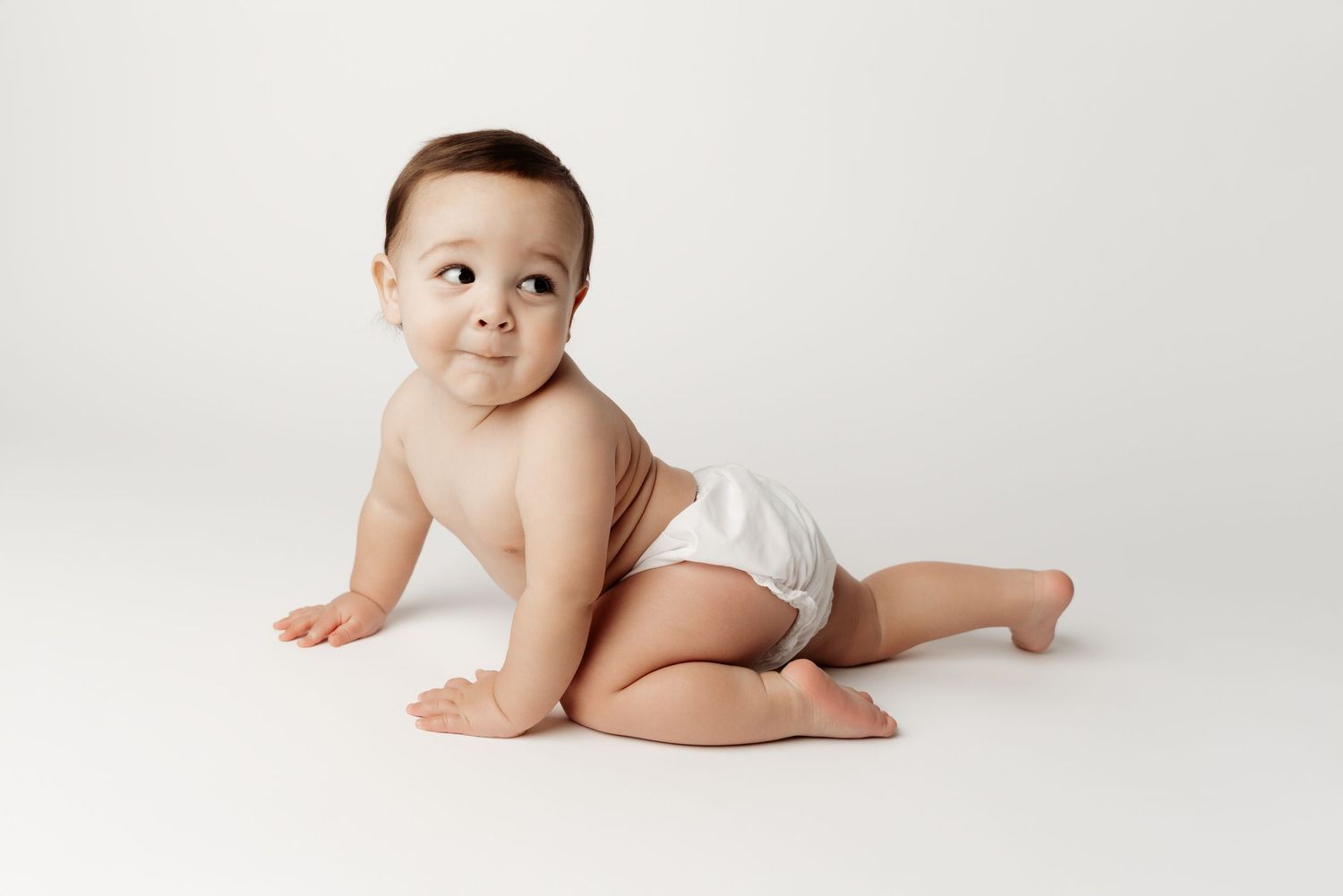 Cute baby in a white diaper crawling on a white background, looking up curiously.