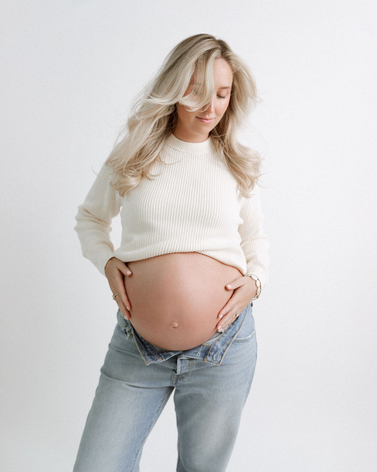 Pregnant woman in white sweater and light jeans shows growing belly in minimalist studio setting.