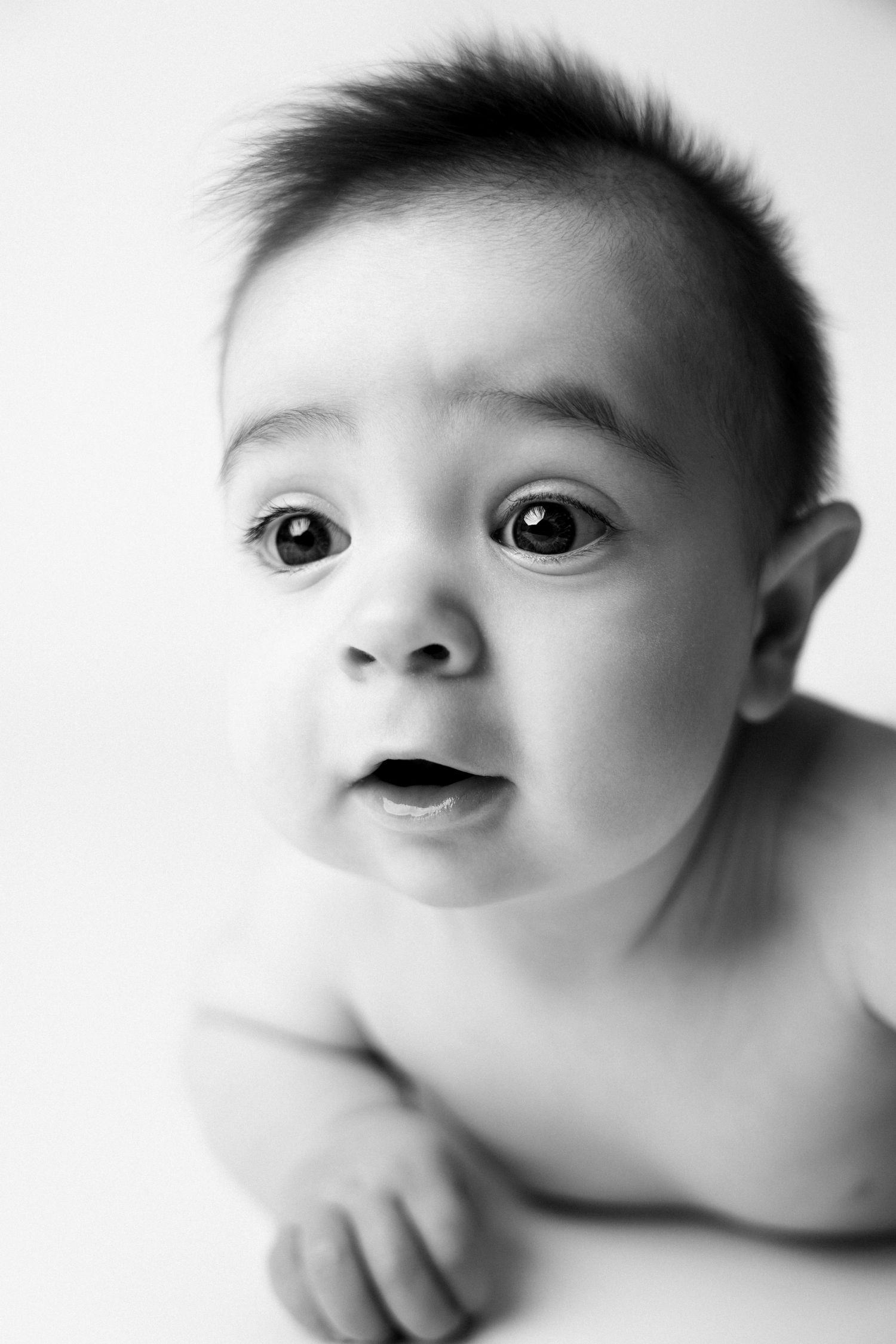 Black and white portrait of an adorable baby with wide eyes lying on stomach, looking up curiously.