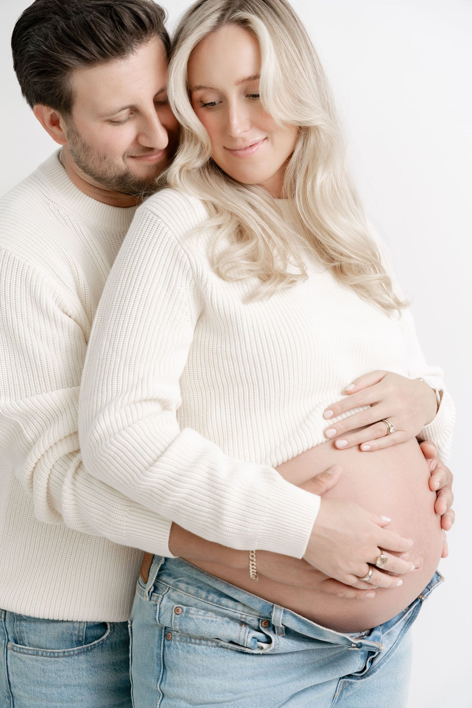 Intimate maternity photos of couple in white sweater and jeans embracing against white background.