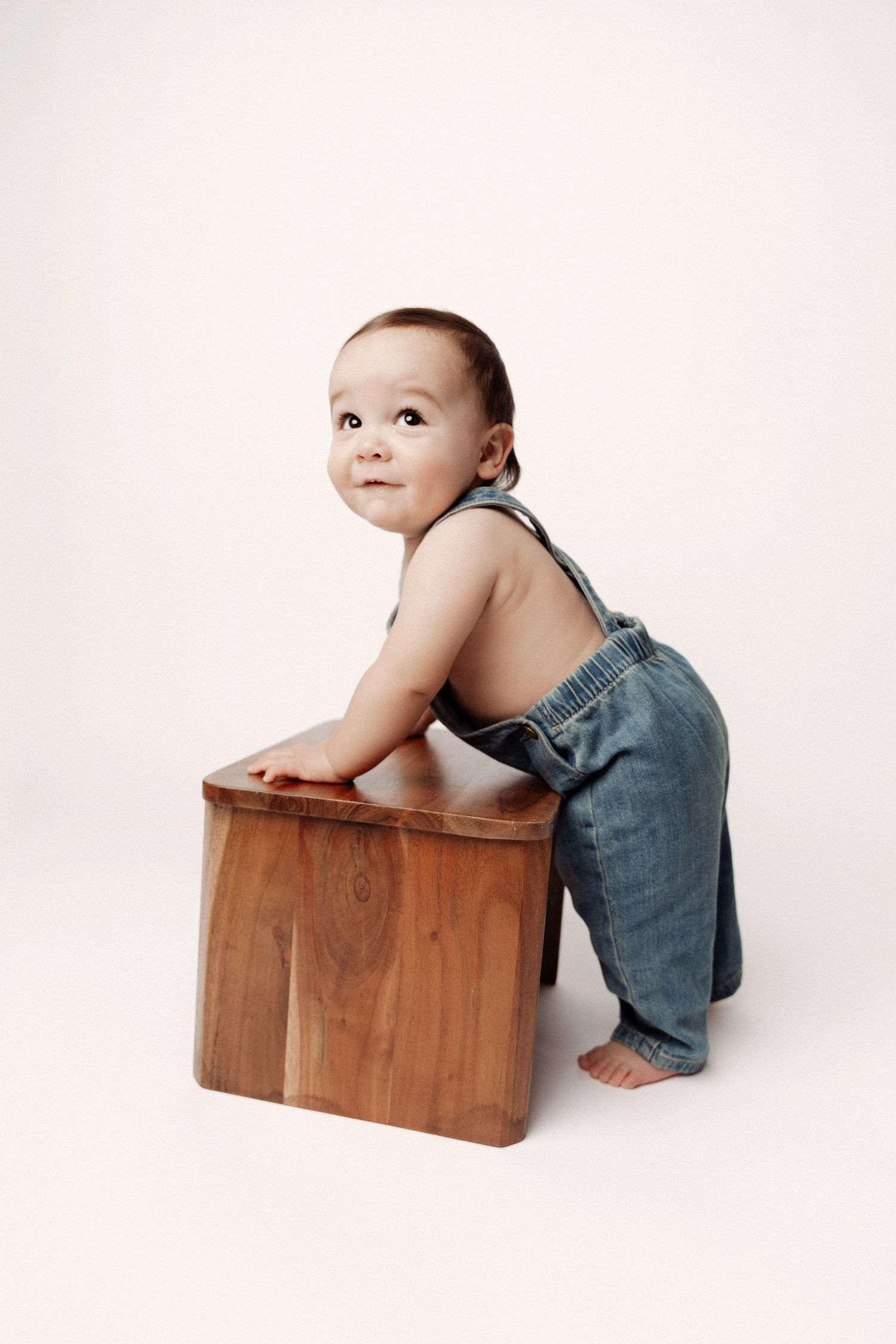 A baby in denim overalls leans against a wooden box in three sequential poses against a white background.