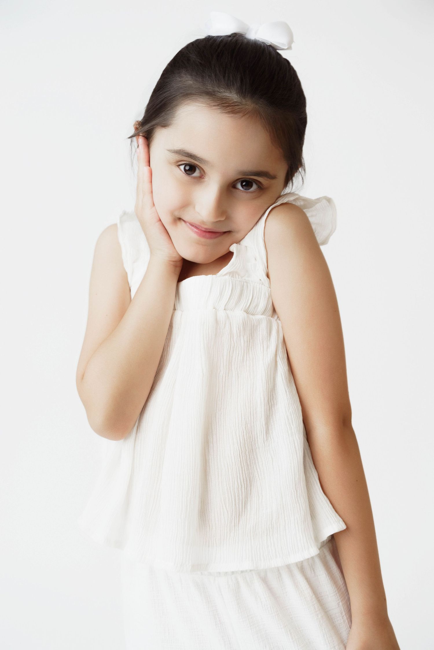 A young girl in a white sleeveless dress smiles softly, resting her hand against her cheek on a white background.