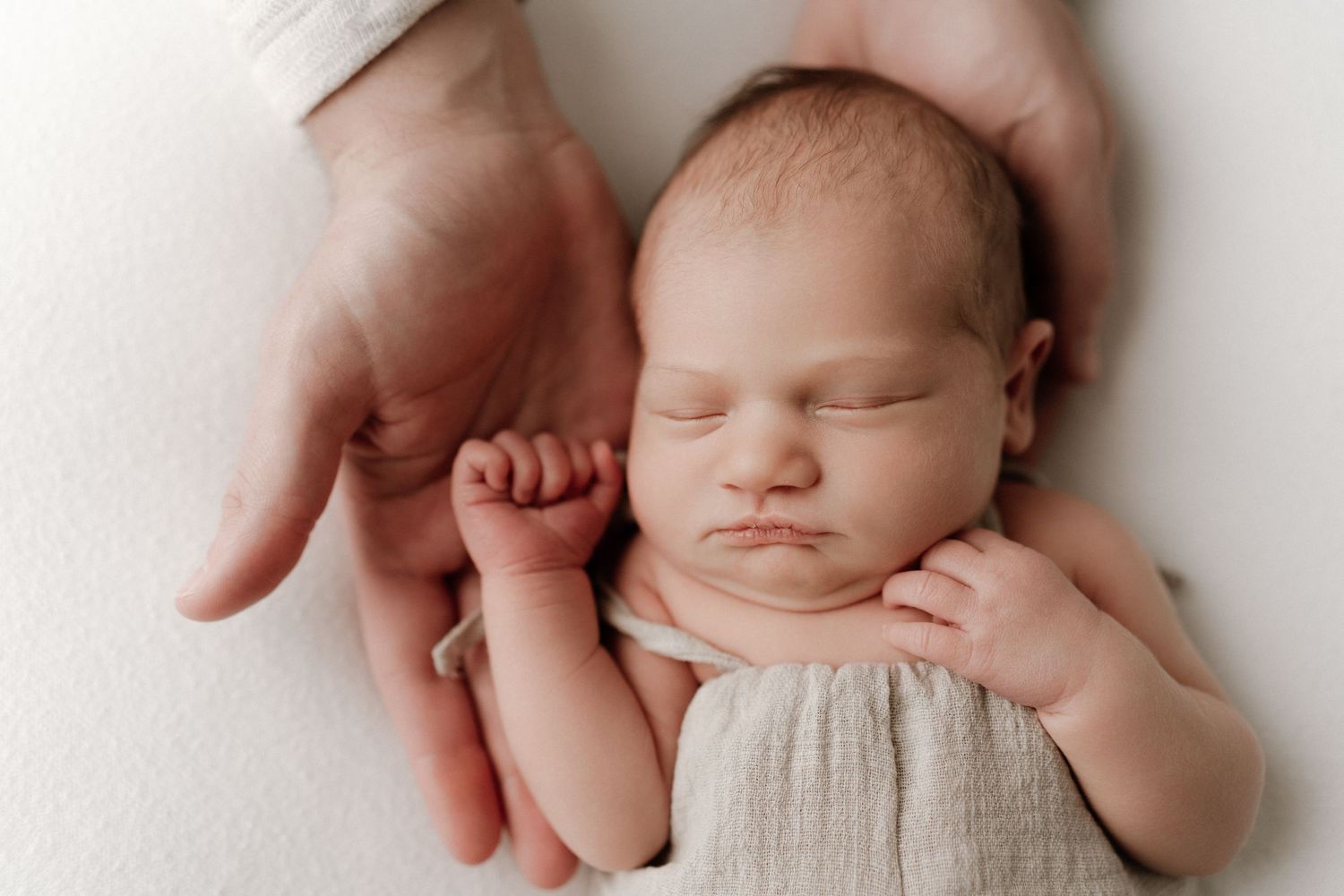 Newborn baby sleeping peacefully in parent's hands, wrapped in a soft beige swaddle on a white background.