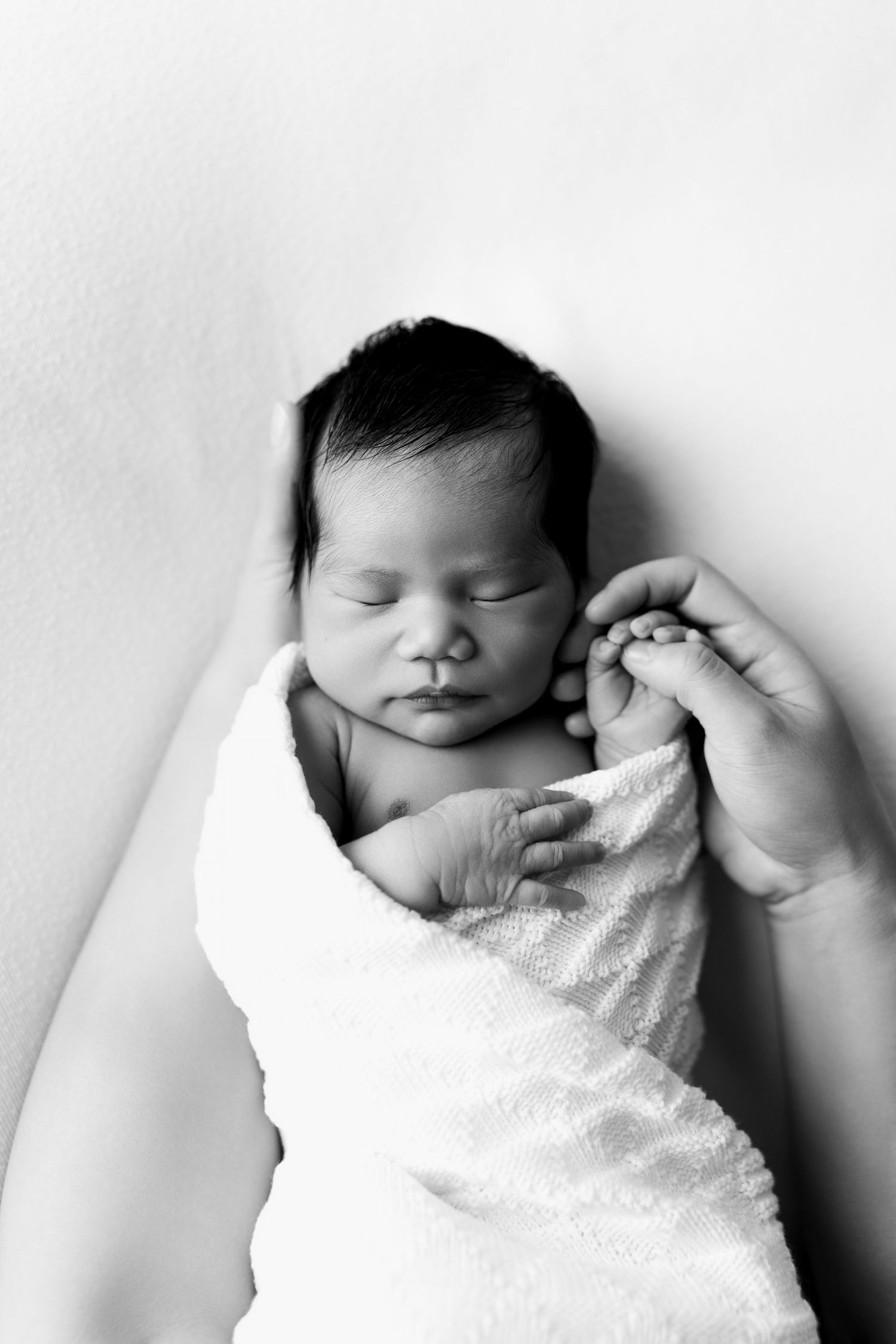 Black and white photo of a sleeping newborn baby swaddled in white wrap, gently held by adult hands.