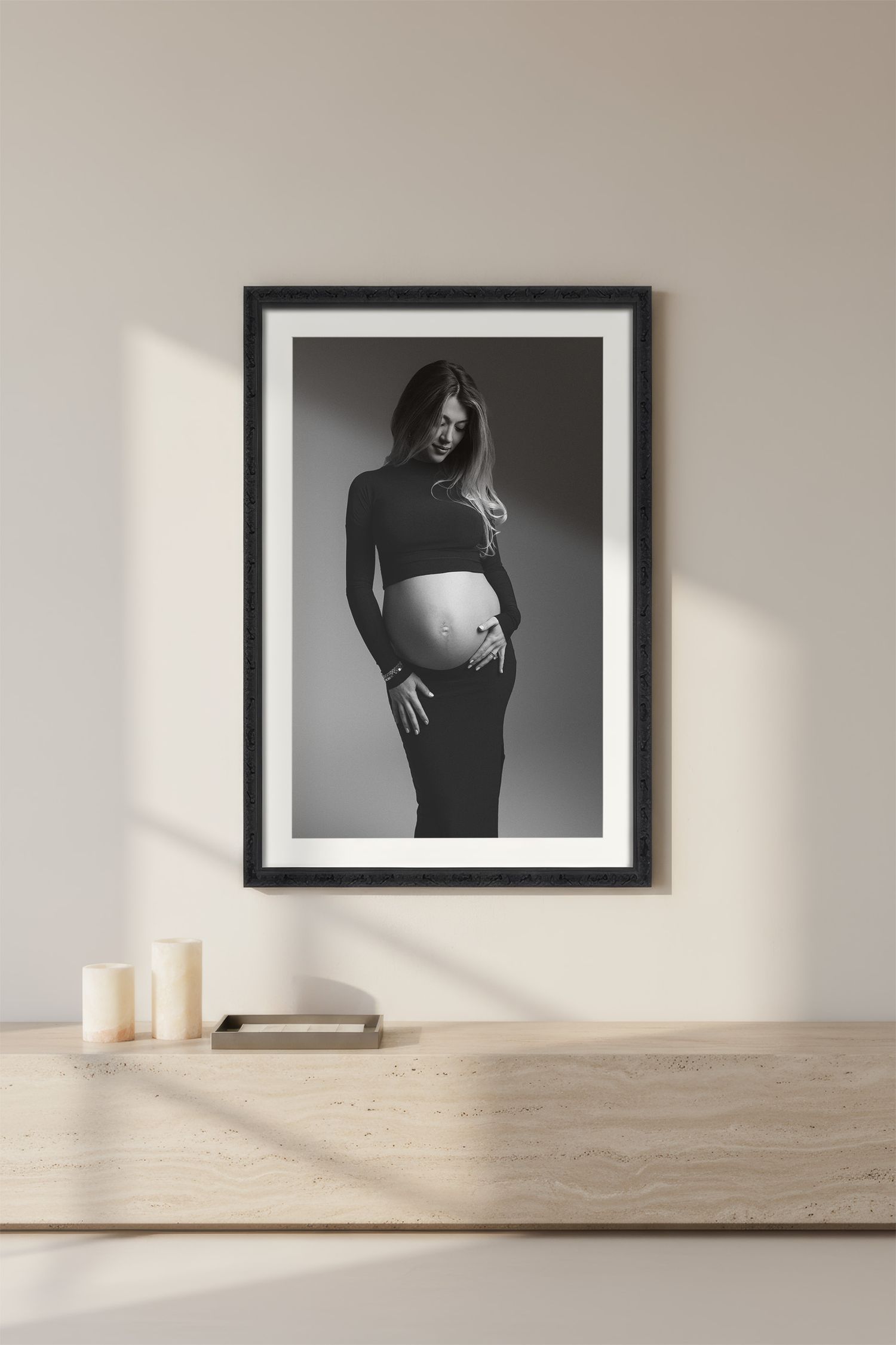 Black and white maternity photography displayed in frames on white wall with natural light and candles on wooden shelf.