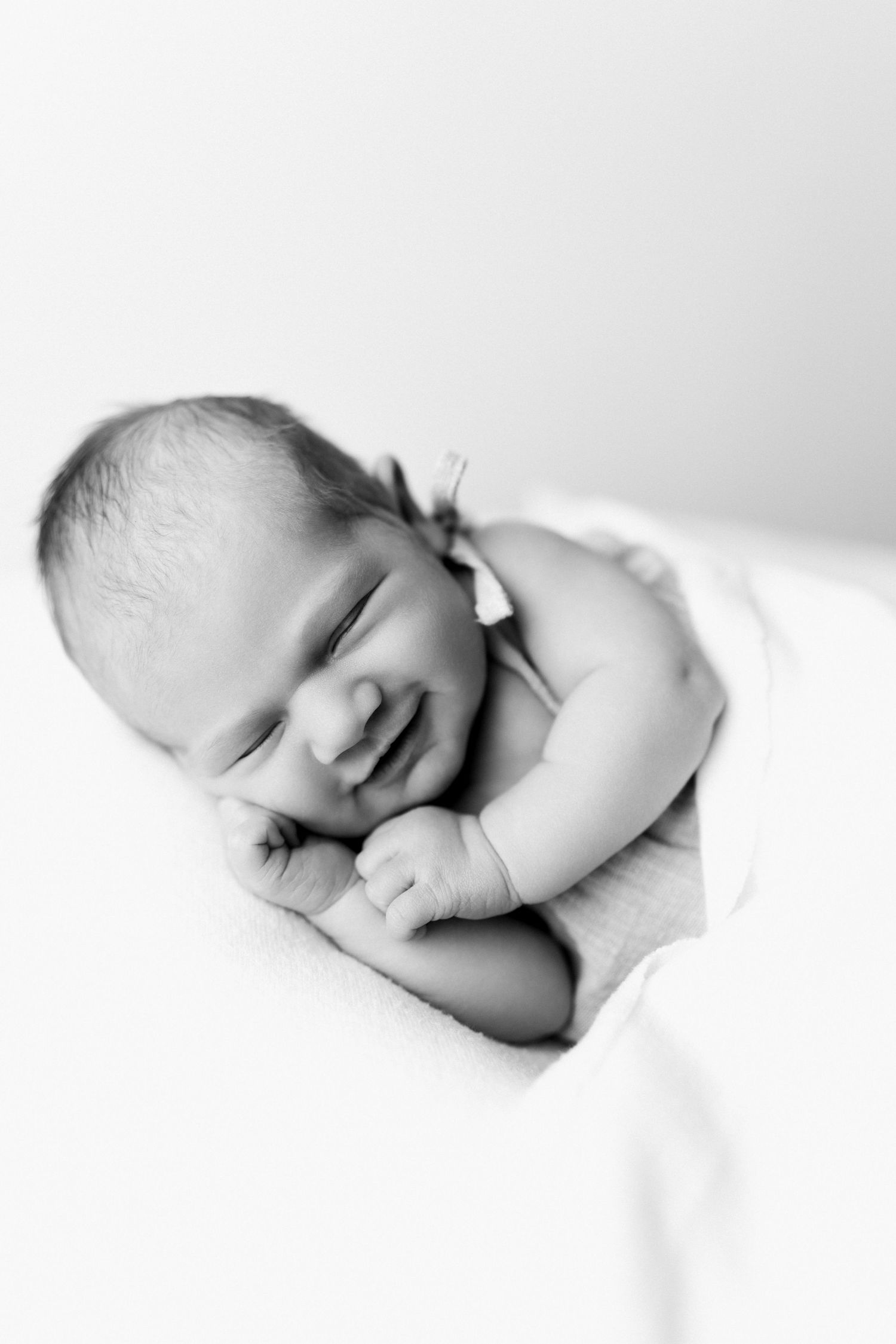 Black and white photo of a smiling newborn baby wrapped in a soft white blanket, peacefully resting.