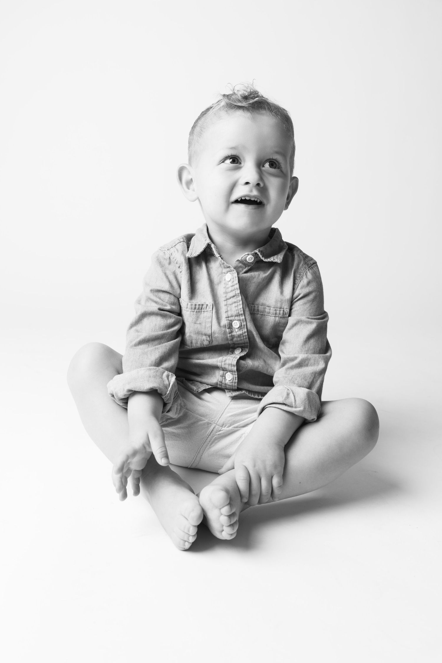 Black and white photo of a happy toddler boy sitting cross-legged, wearing a denim shirt, looking up and smiling.