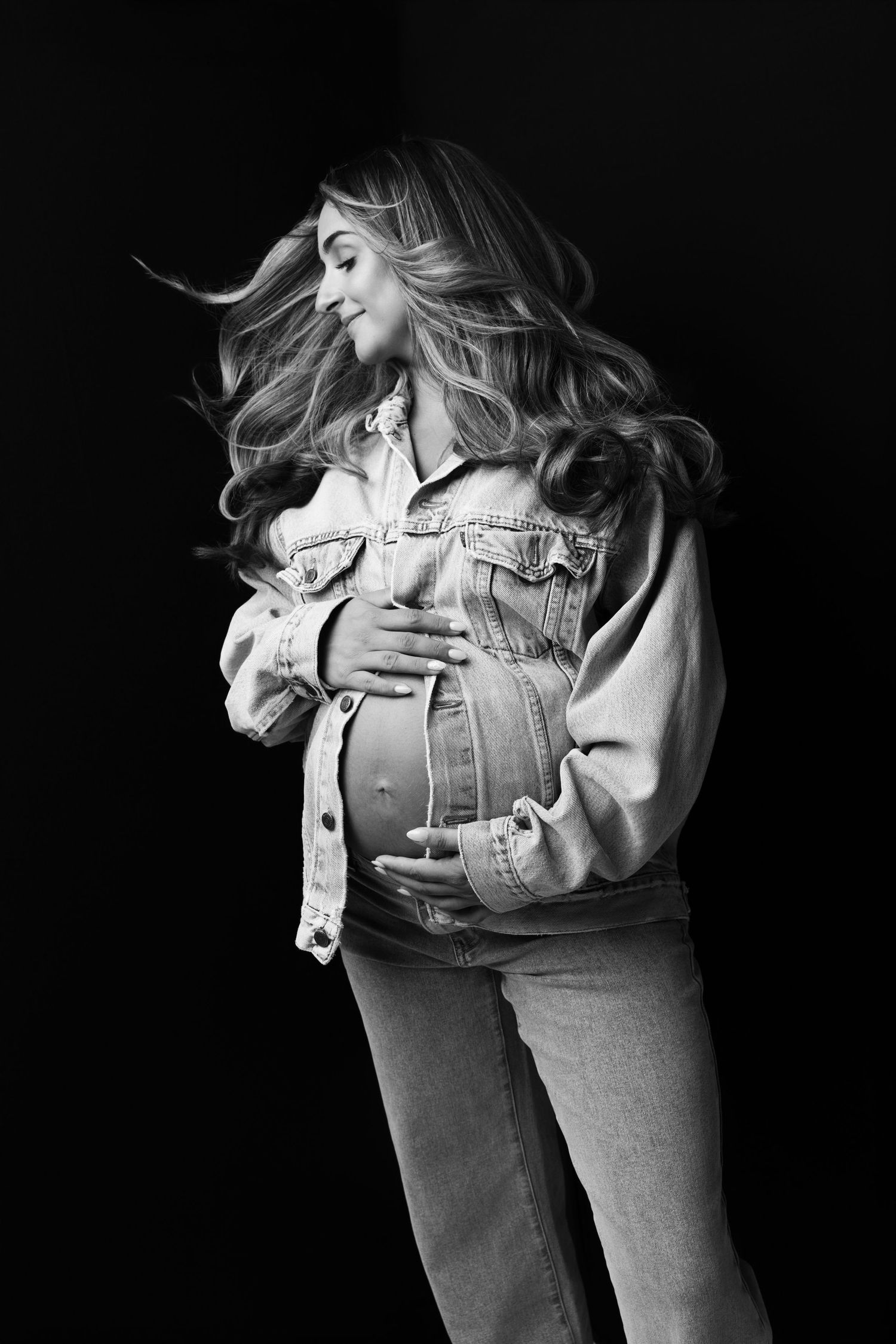 Black and white photo of a pregnant woman in denim jacket cradling her baby bump with flowing hair against dark background.