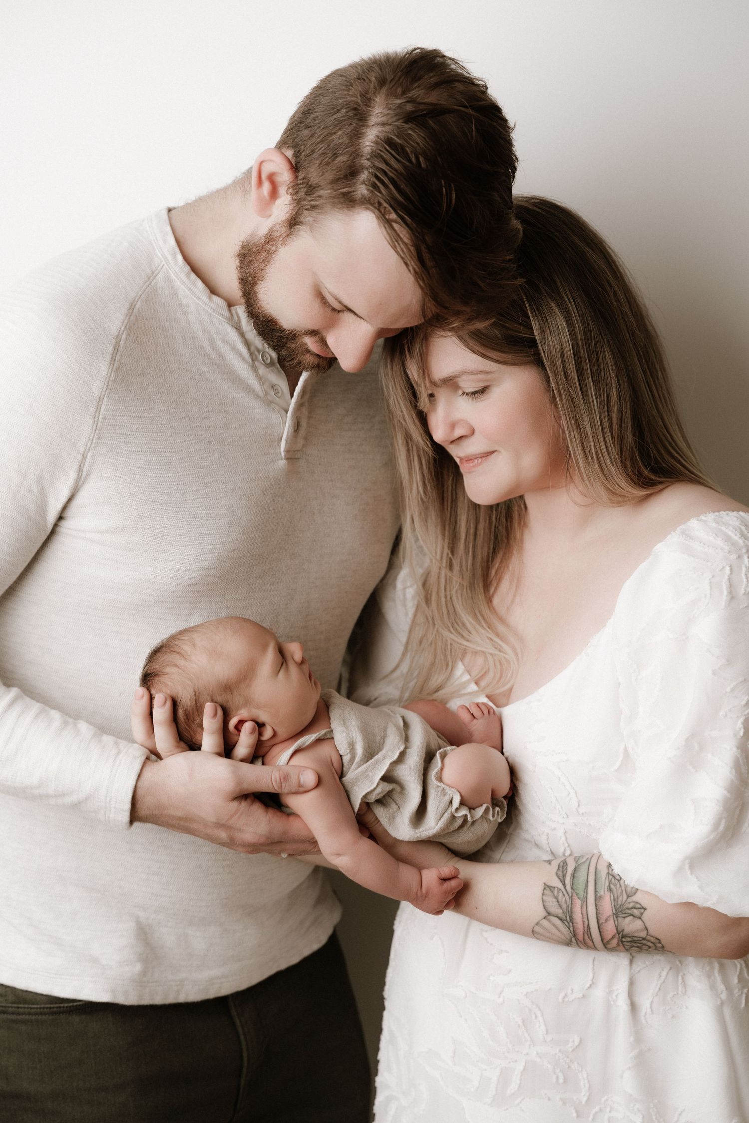 New parents tenderly cradling their sleeping newborn baby together in a soft, neutral-toned studio portrait.