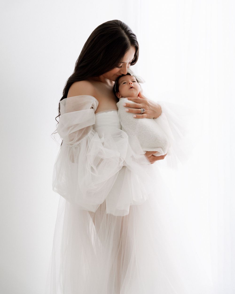 A mother in a white tulle gown tenderly kisses her newborn baby during an elegant studio portrait session by a Michigan family photographer