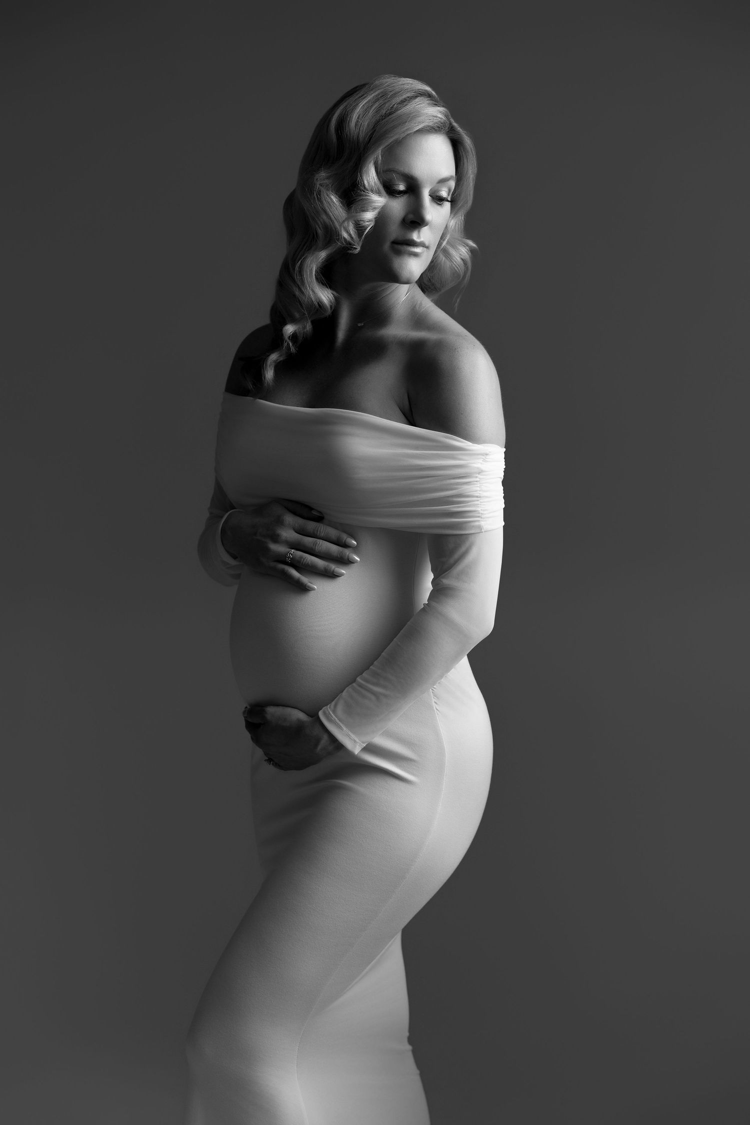 Elegant maternity portraits in off-shoulder white dress with dramatic studio lighting.