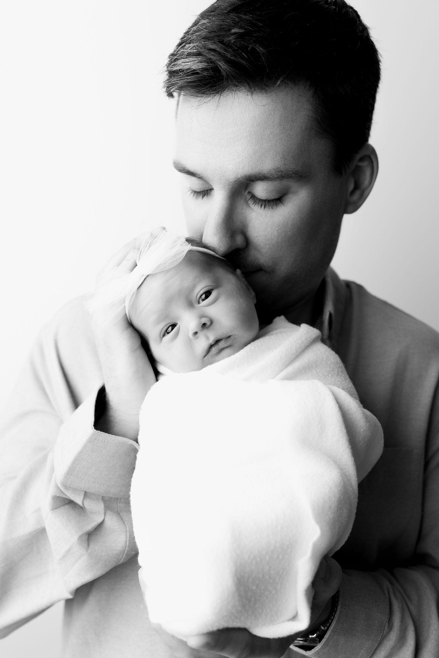 Black and white photo of a father tenderly kissing his swaddled newborn baby on the forehead.