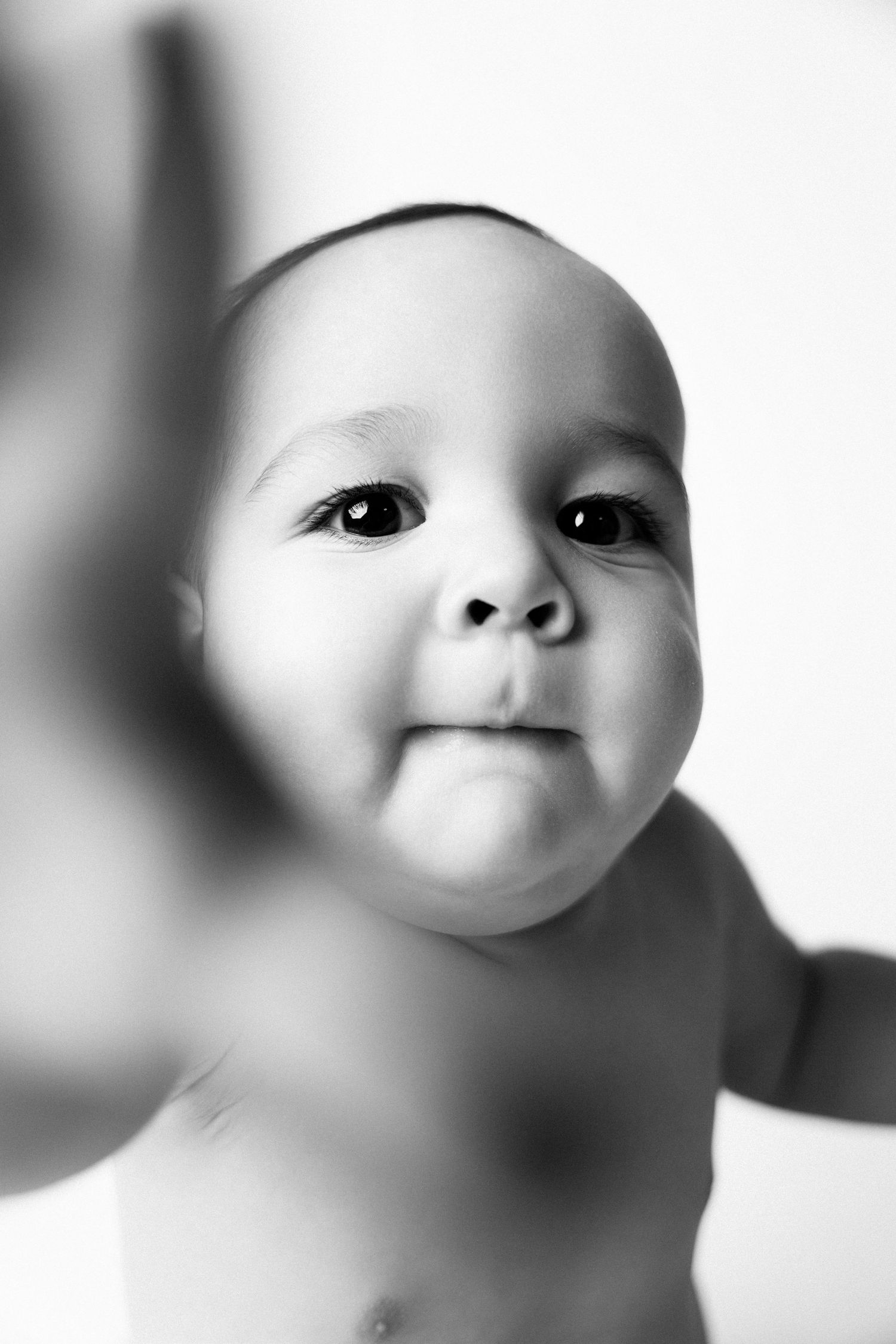 Black and white portrait of an adorable baby reaching toward camera with curious wide eyes and chubby cheeks.