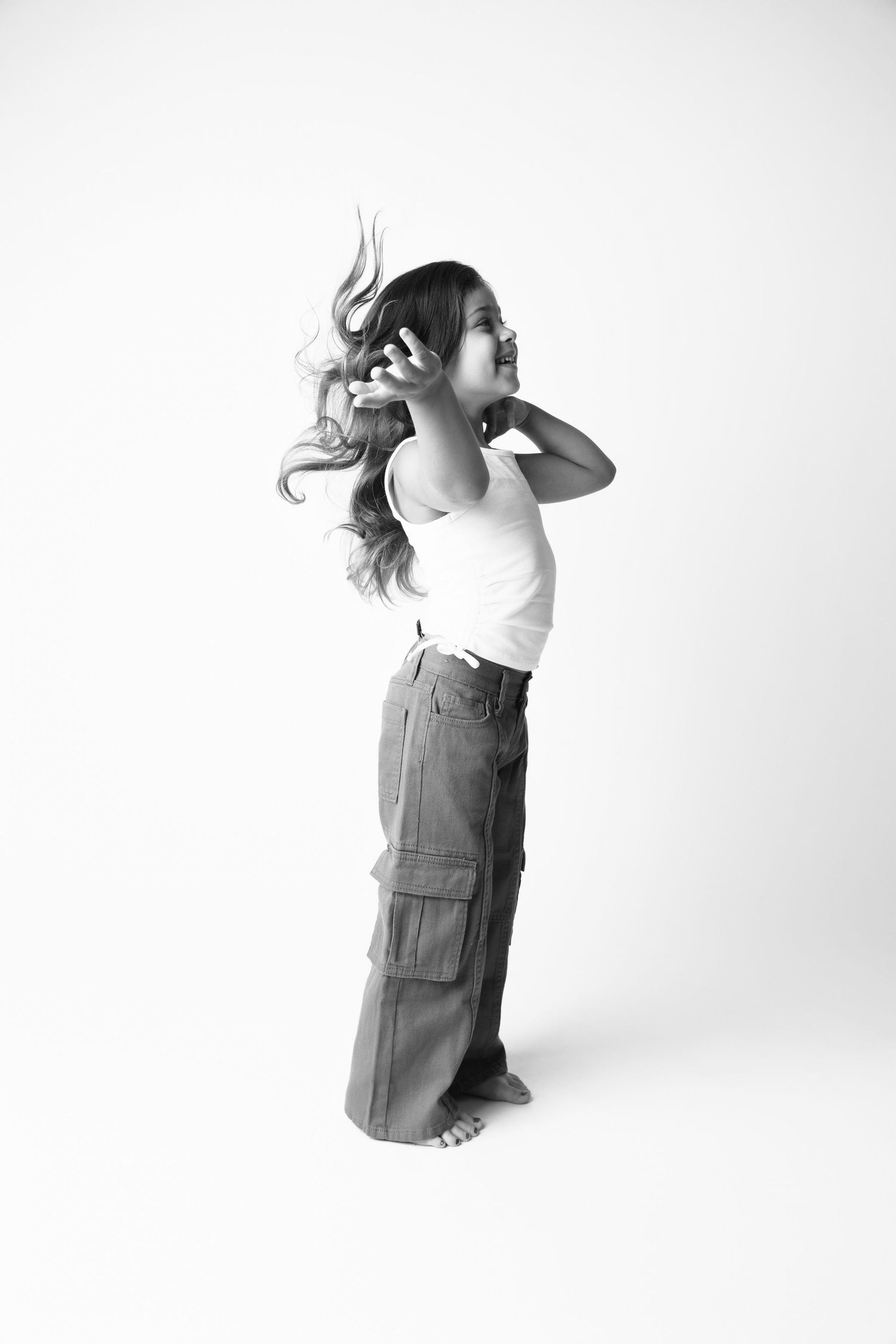 Black and white photo of a young girl tossing her long hair, wearing white tank top and wide-leg cargo pants.