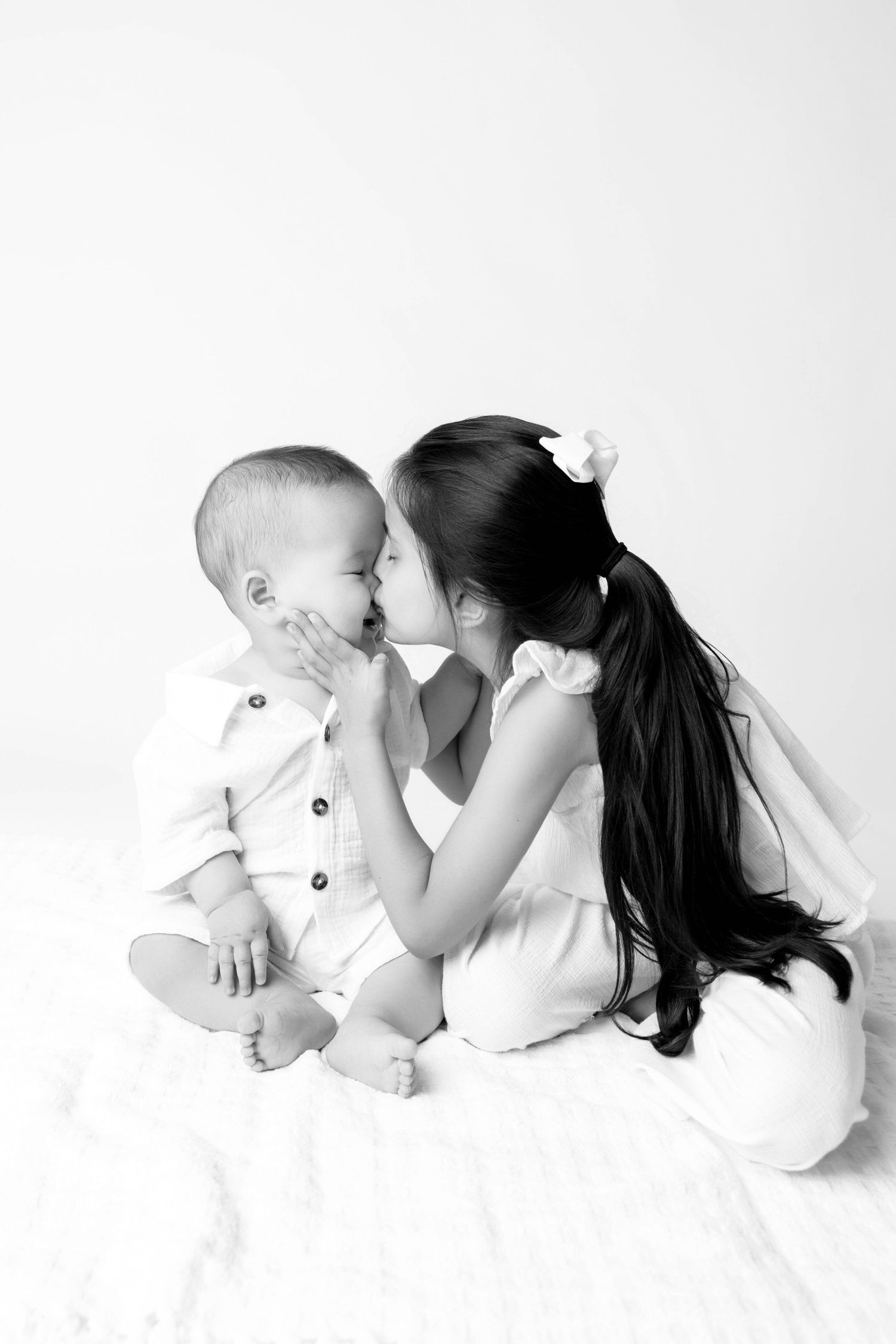 Black and white photo of a young girl with long hair kissing a baby sibling on the cheek while sitting together.