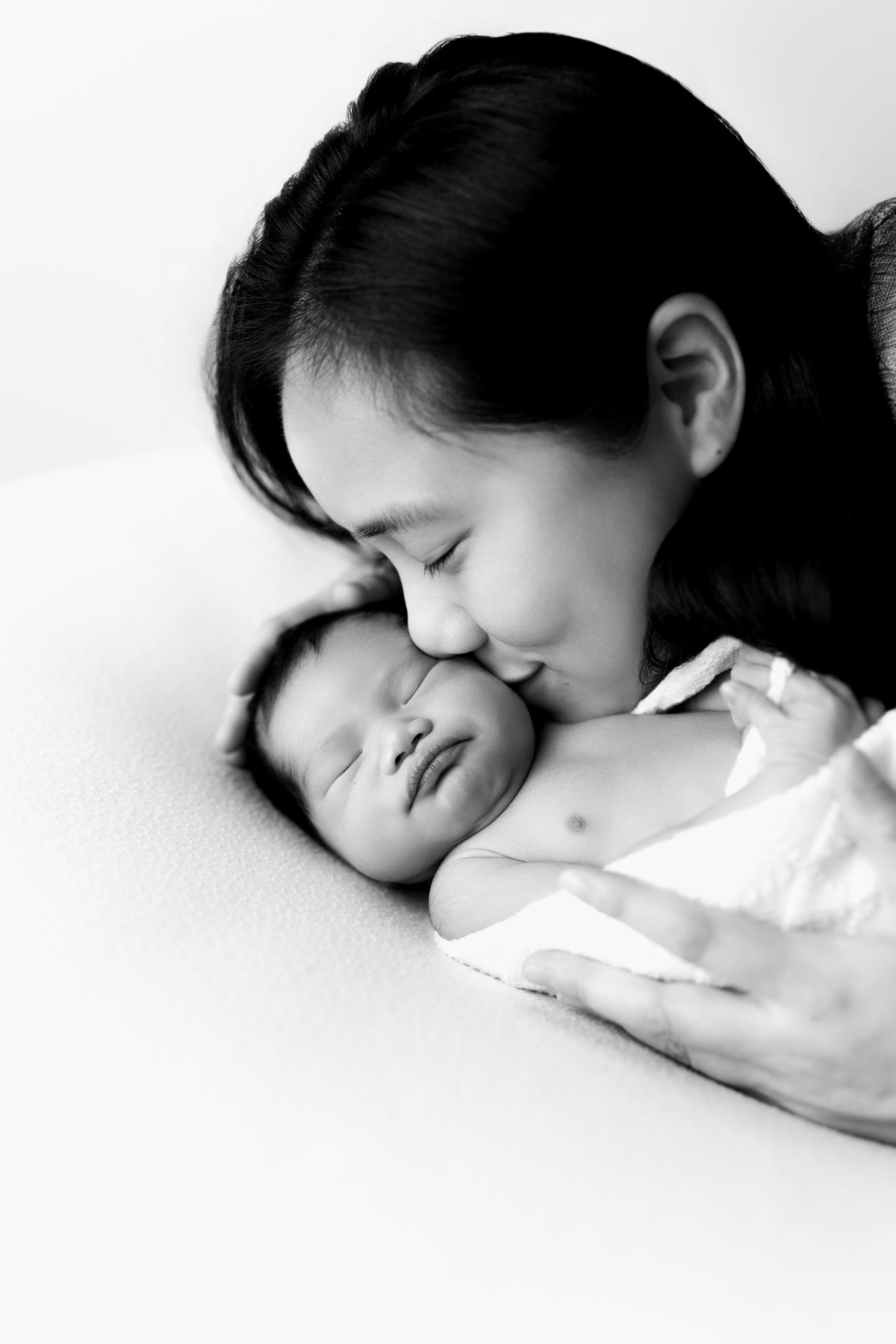 Black and white photo of a mother tenderly kissing her sleeping newborn baby wrapped in white cloth.