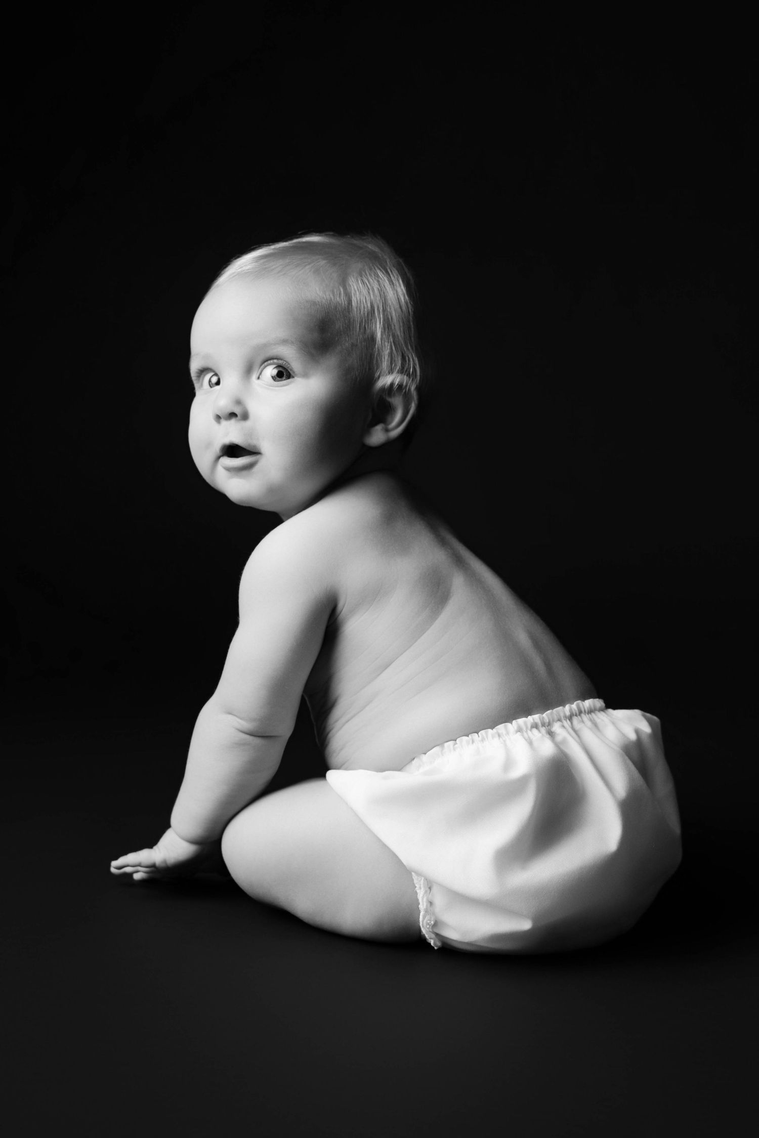 Black and white portrait of a cute baby sitting in a diaper against a dark background, looking over shoulder.