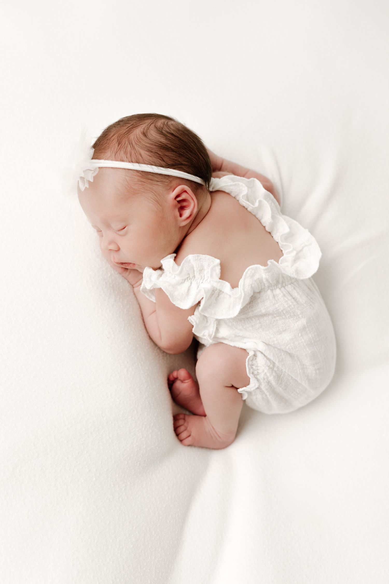 Peaceful newborn baby sleeping in white ruffled romper and headband on soft white blanket.