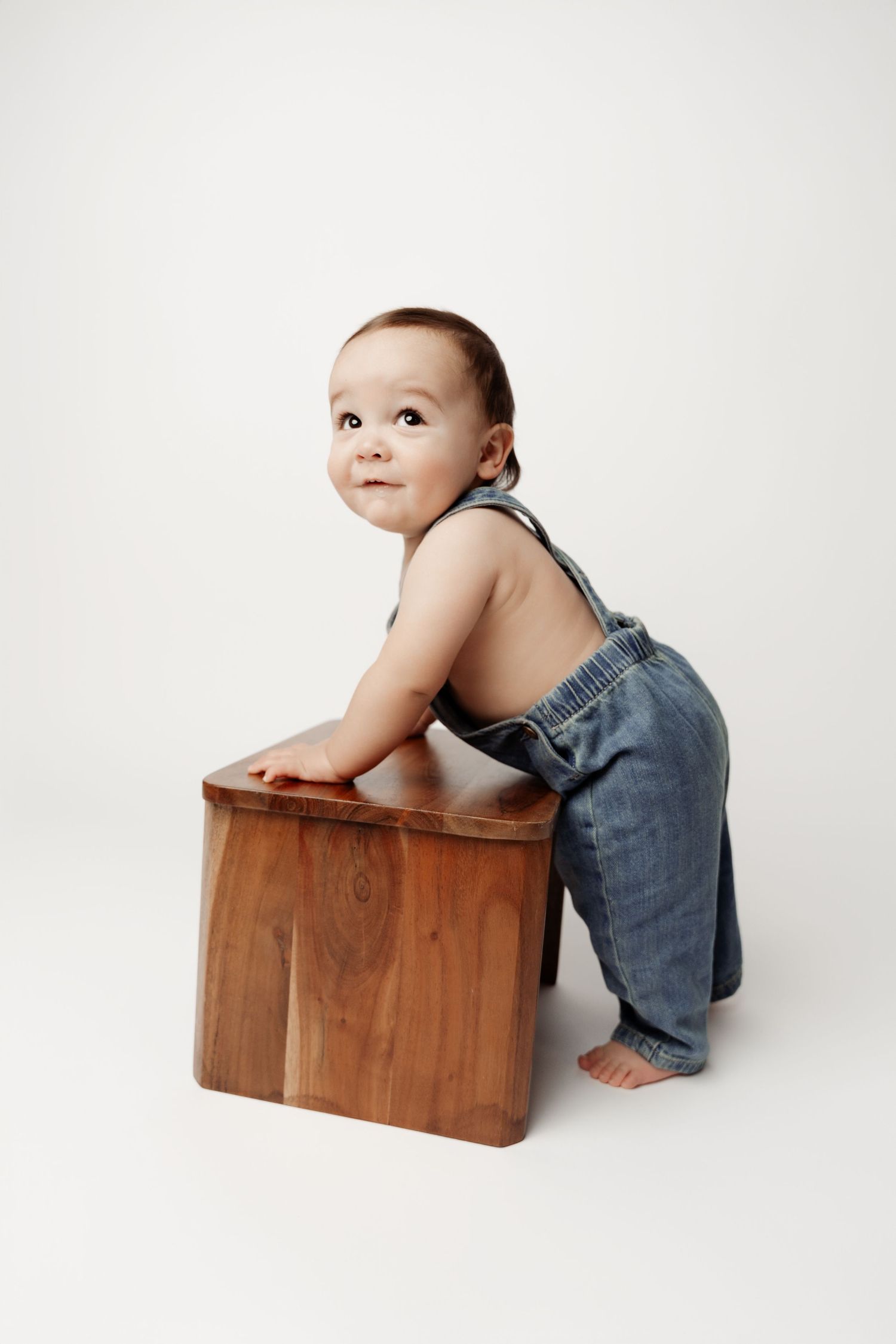 Adorable baby in denim overalls leaning on a wooden cube, looking up curiously against a white background.