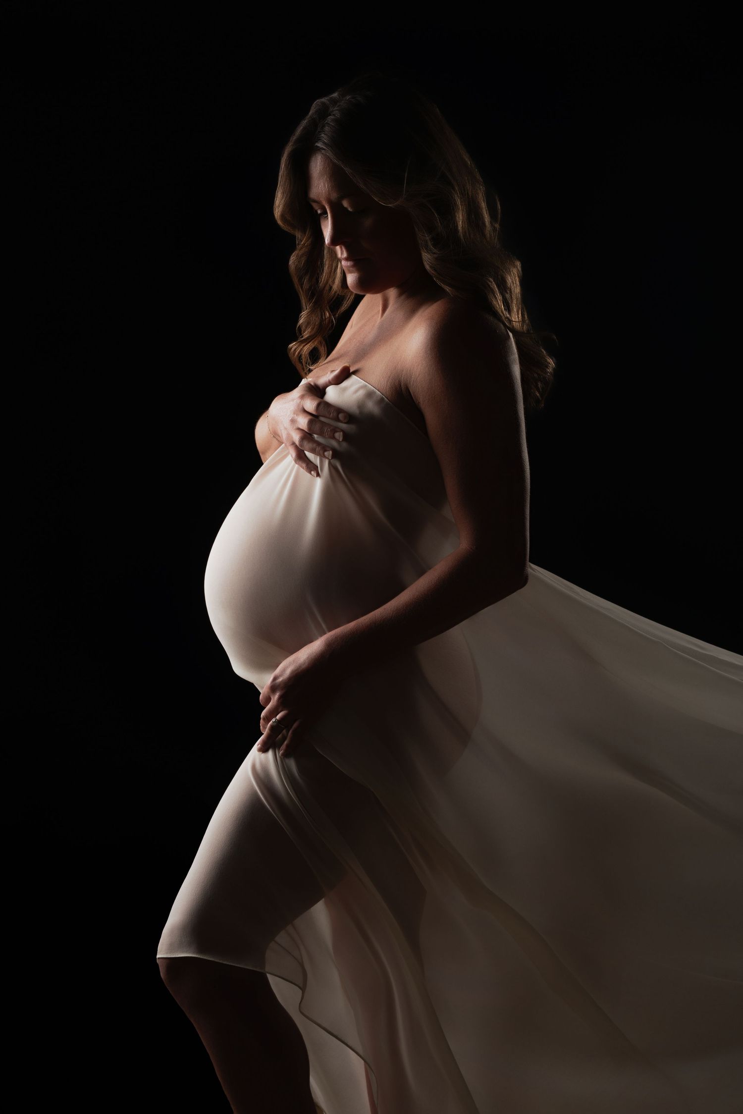 Elegant pregnant woman in flowing white fabric gown posing gracefully against a dark black background in a maternity portrait.