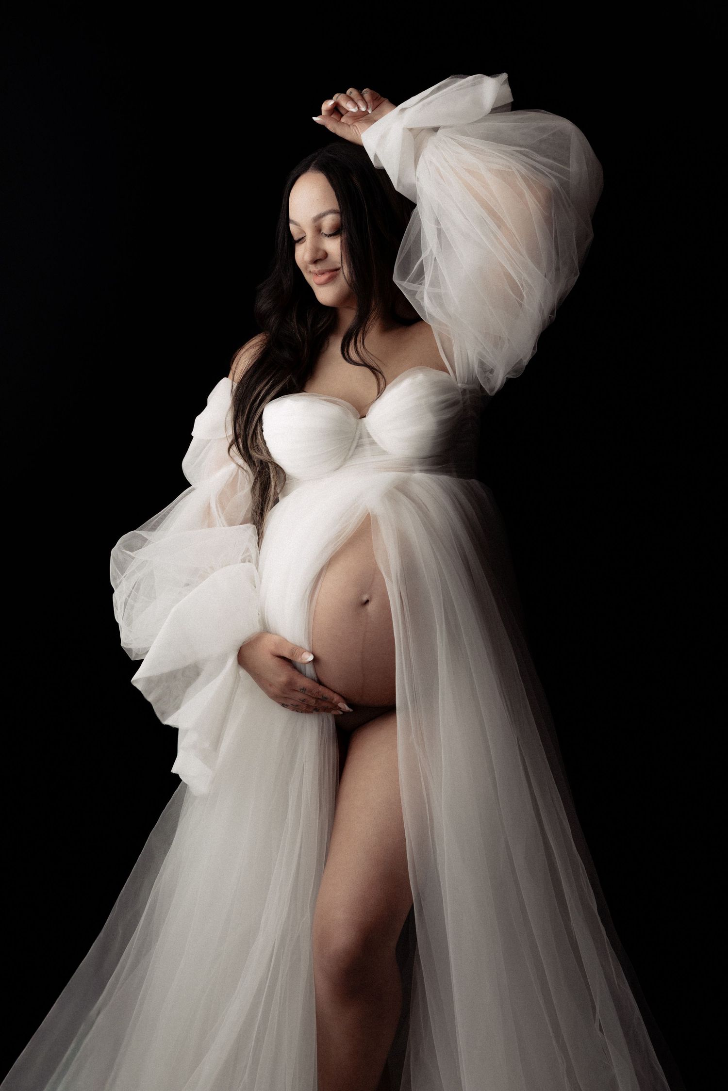 Pregnant woman in flowing white tulle gown cradling baby bump against black background in elegant maternity photo.