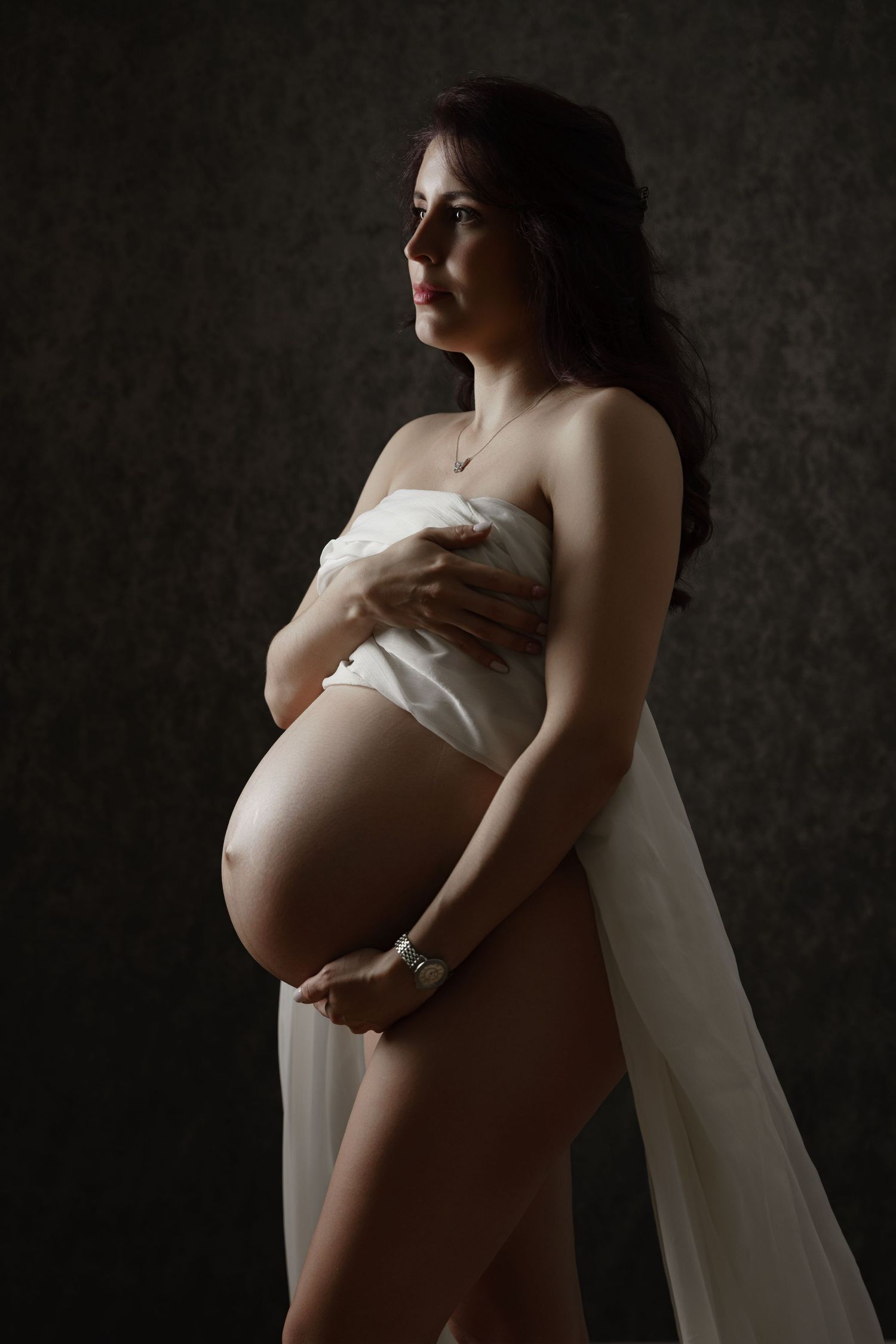 Dramatic maternity photography with woman in white fabric against dark background.