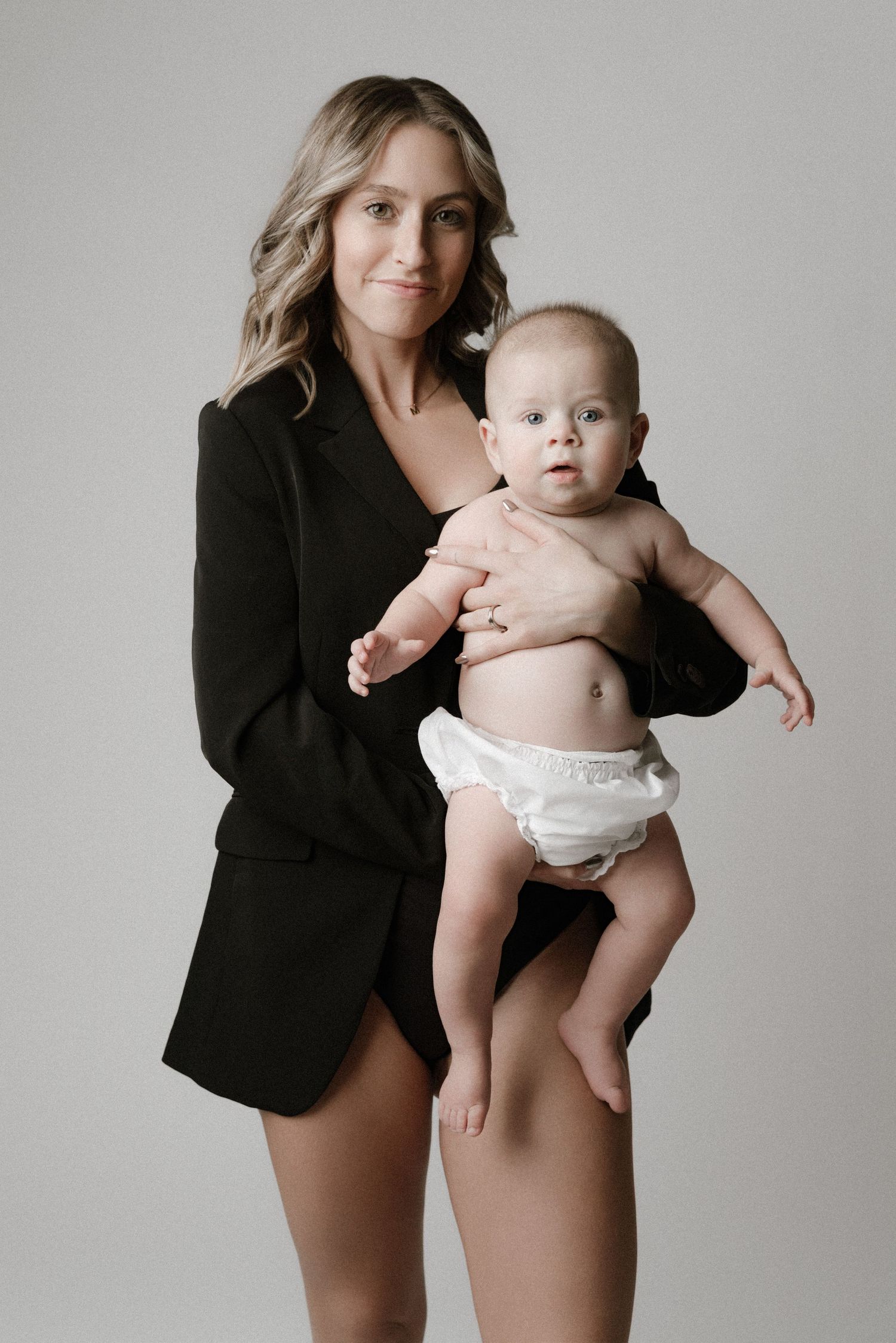 Studio portrait of parent in black blazer holding baby in white diaper against neutral gray background.