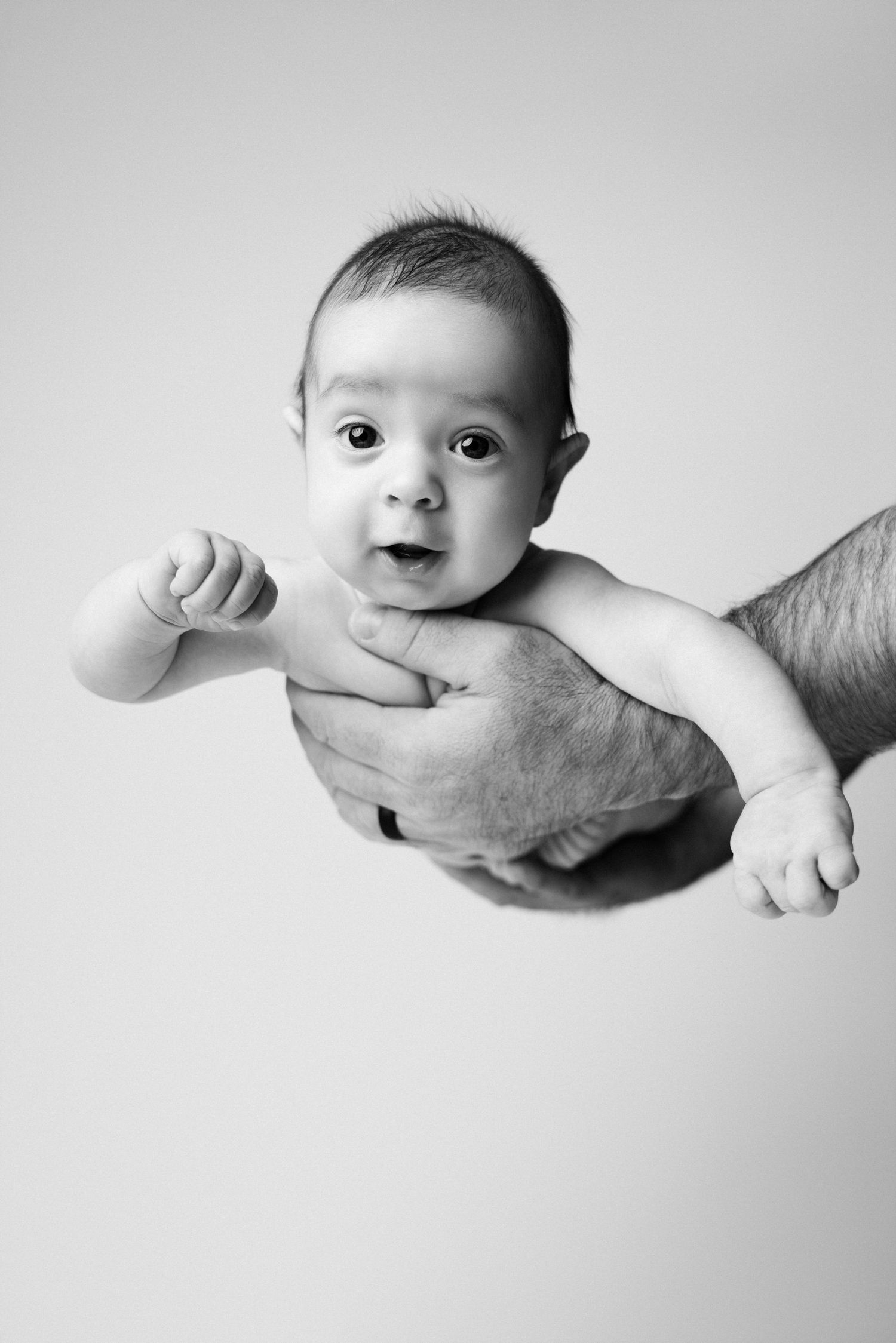 Black and white photo of a newborn baby held in adult hands, looking alert with wide eyes and open mouth.