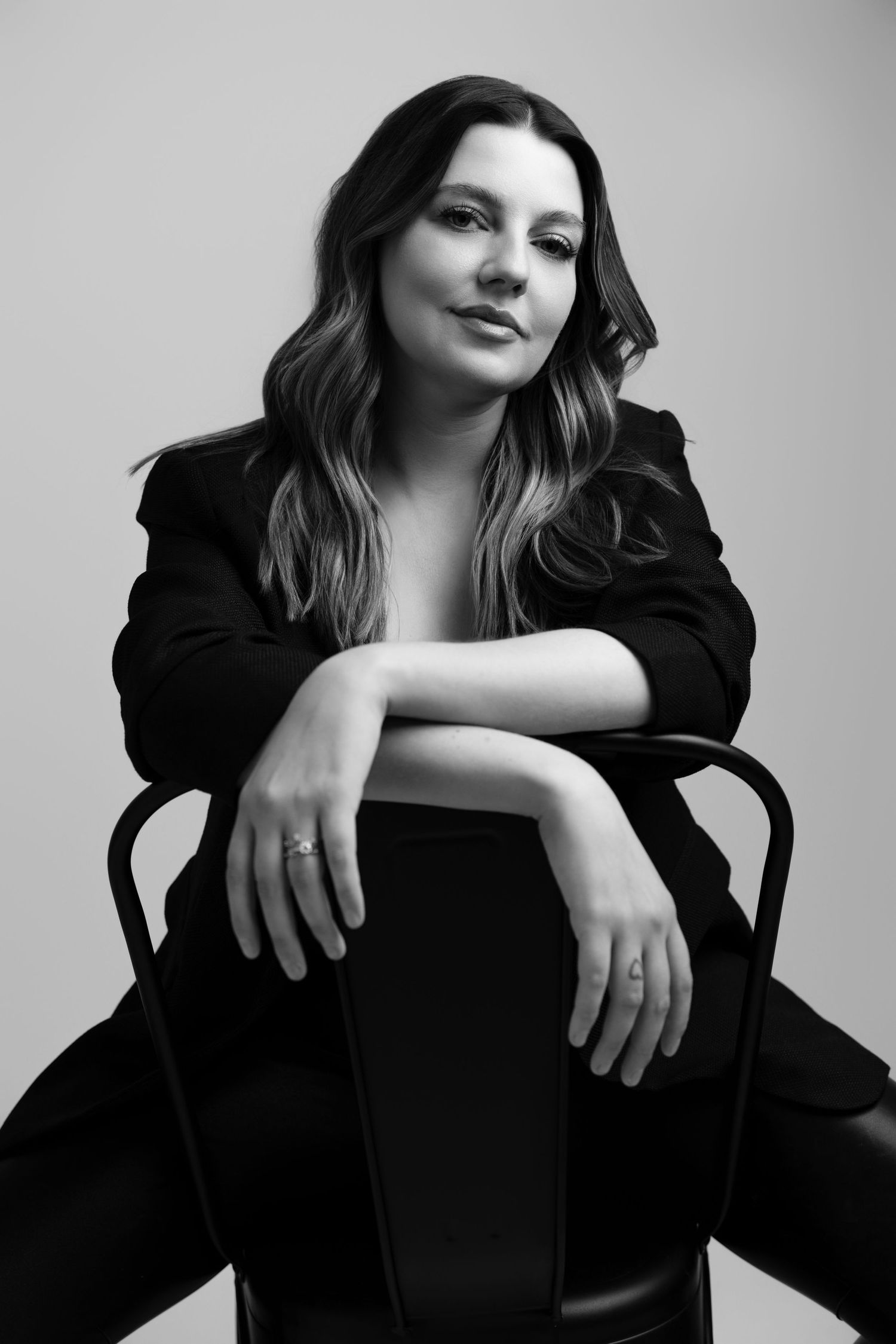 Black and white headshot of a woman in a black blazer sitting on a chair, posing confidently in a studio in Michigan.