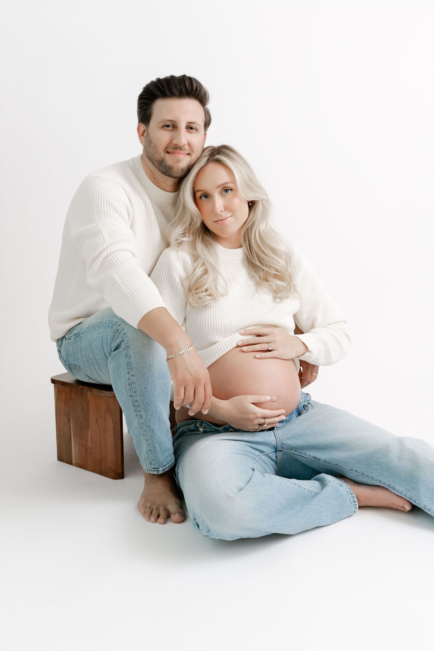 A couple in matching white sweaters and jeans pose together for a maternity photo against a white background.